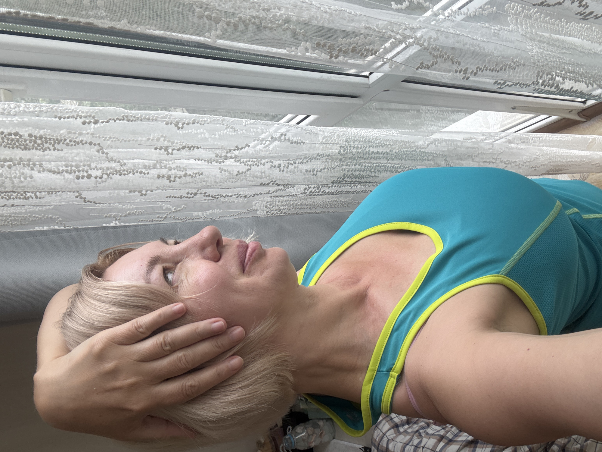 The image shows a woman with short blonde hair, wearing a turquoise tank top with lime green trim. She is looking out of a window with white lace curtains. Her left arm is bent with her hand resting on the back of her head. The scene appears to be indoors, with a bed visible in the background to the left. The presence of lace curtains and a window suggests an interior setting, possibly a room in an apartment or hotel. The lighting indicates it is daytime. There is no visual information in the image that can identify the location as Sochi, Russia.
