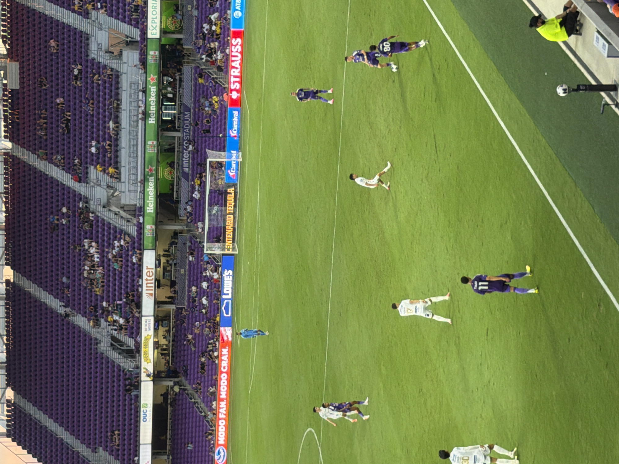 This image captures a soccer match at Inter&Co Stadium in Orlando, Florida. The stadium is filled with purple seating, most of which is unoccupied. A soccer field with green grass and white lines is visible in the foreground. Several soccer players are scattered across the field, some in white uniforms and others in purple. Advertisements for Lowe's, Heineken, and Carnival are displayed on the stadium walls. In the lower right corner, a person in a neon yellow shirt is seated. The overall scene suggests a sporting event in progress, with the stadium partially filled.