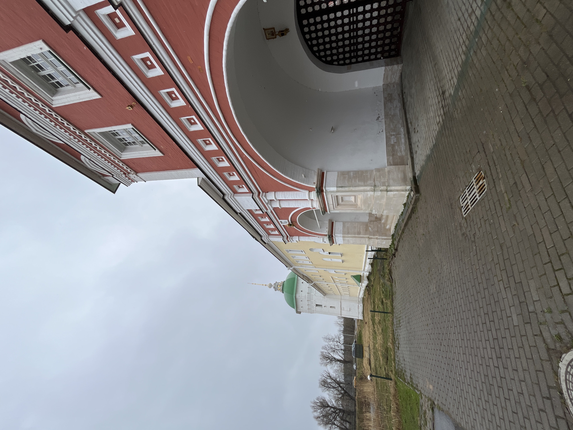 The image depicts a view of a historical architectural structure with red, white, and light-yellow facades. The architectural details include arched openings, windows, and decorative elements. The foreground consists of a brick-paved path. In the background, there is a green-domed building and some bare trees, with an overcast sky above. The photograph appears to be taken in Луговой, Russia.