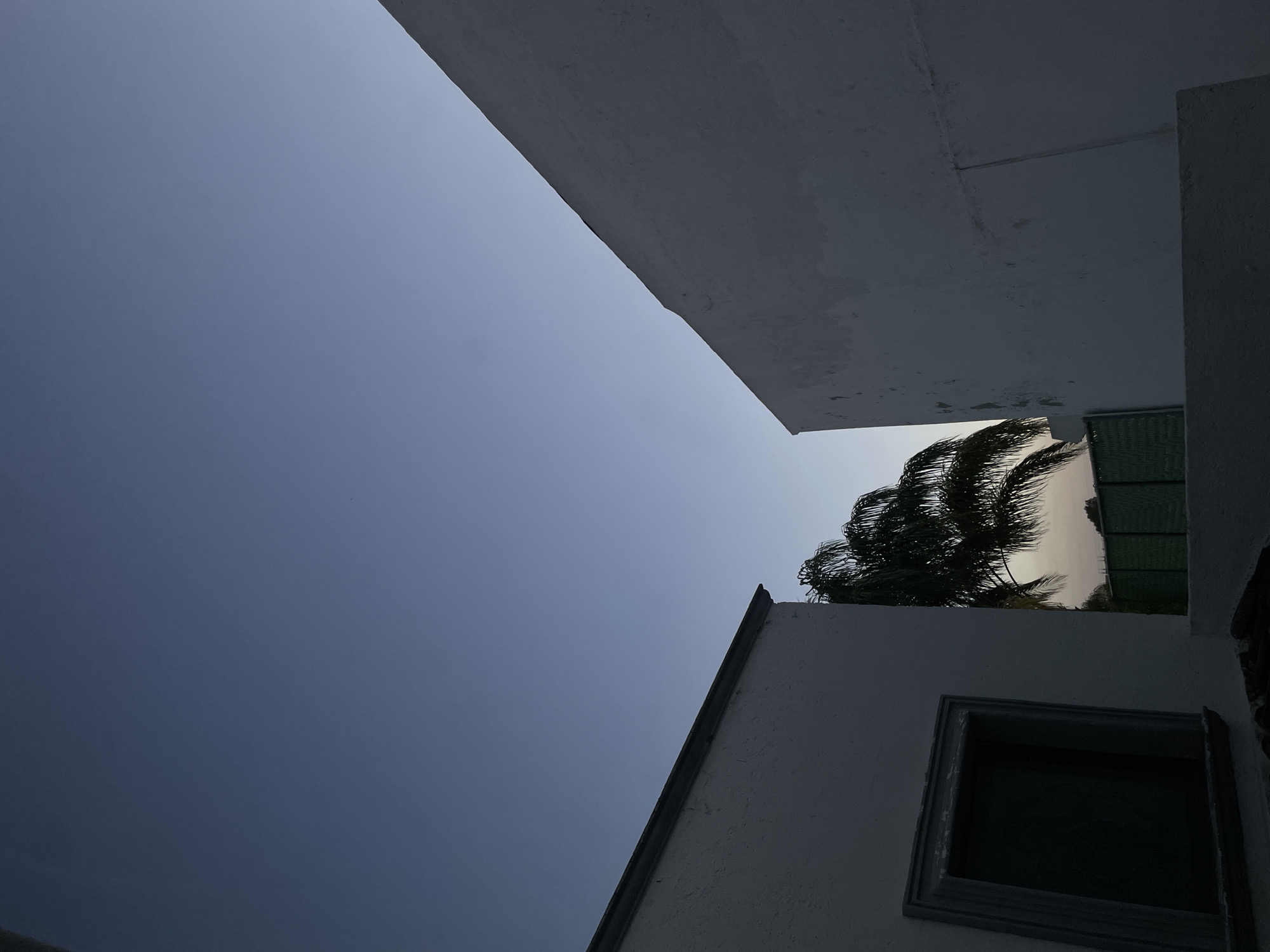 A low-angle shot reveals a section of the sky framed by architectural structures, likely taken in San Bernardino Tlaxcalancingo, Mexico. The sky transitions from a light blue to a darker hue. White buildings with sharp edges and flat surfaces dominate the frame. A palm tree and a green grate are visible. A window frame, without glass, is present on one of the buildings. The overall scene is bathed in shadows.
