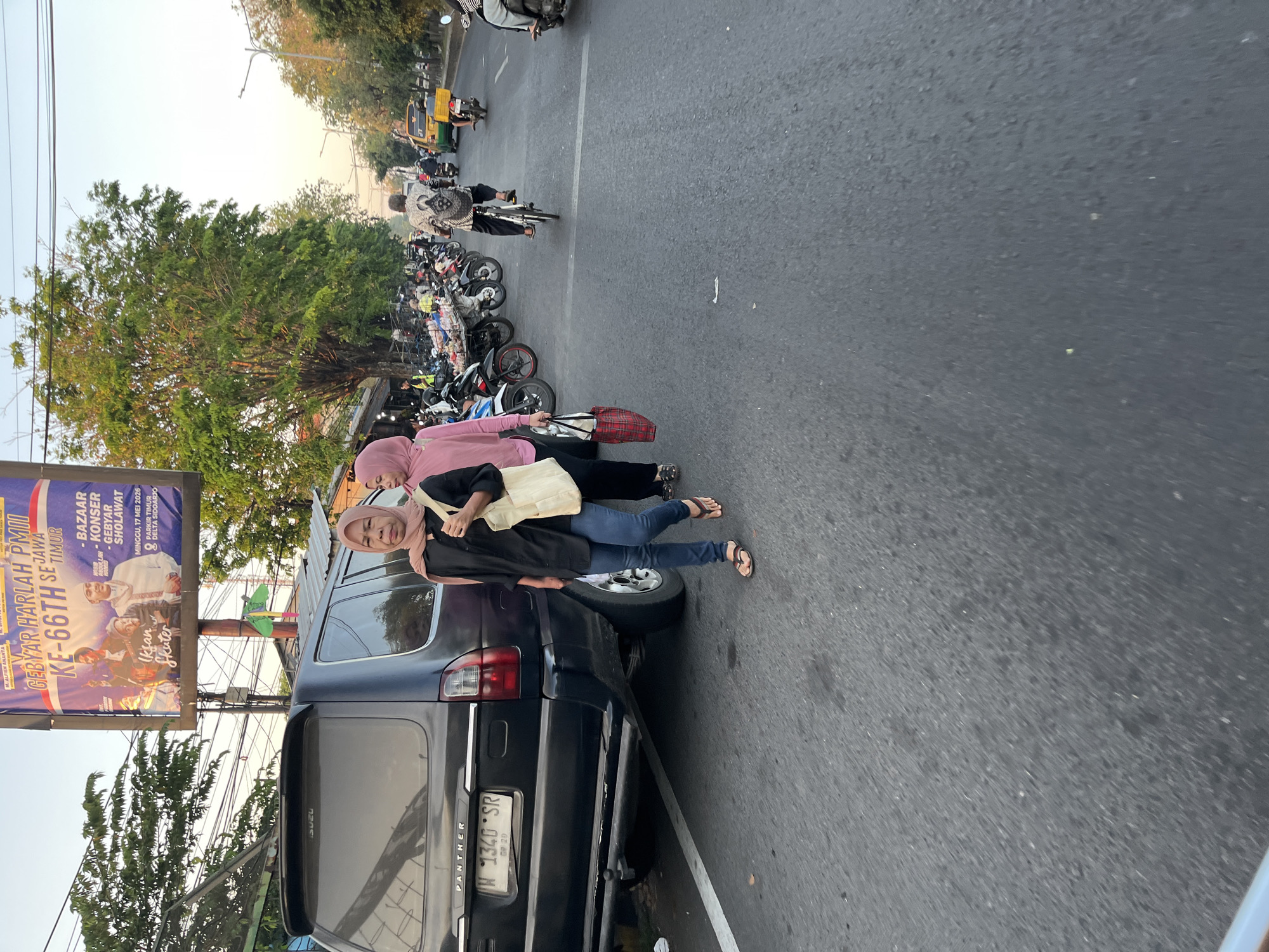 A street scene in Ngaban, Indonesia, captures a moment at dusk. In the foreground, a black SUV is parked on the side of the road, its rear end visible. To the right of the SUV, two women wearing hijabs are walking on the pavement. The woman closer to the camera wears a black shirt and blue jeans, carrying a light-colored tote bag over her shoulder. The second woman, further from the camera, wears a pink shirt and is also carrying a tote bag.
The street is lined with parked motorcycles and vendor stalls, suggesting a busy marketplace. Beyond the stalls, a row of trees with lush green foliage lines the street. In the background, a billboard promotes an event labeled "GEBYAR HARLAH PMII KE-66TH SE JAWA TIMUR" and mentions "BAZAAR - KONSER - GEBYAR SHOLAWAT". Traffic is visible on the road, including motorcycles and a yellow three-wheeled vehicle. The sky is a light orange-pink, indicating the setting sun. The overall atmosphere is that of a vibrant, active urban environment during an evening gathering.