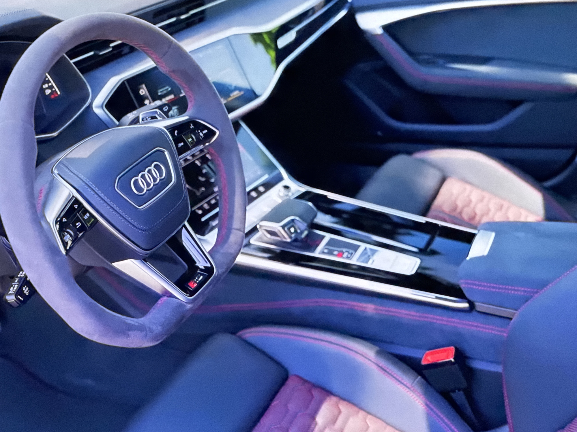 The image shows the interior of a car, likely an Audi, with a focus on the steering wheel and center console. The steering wheel is covered in a textured material, possibly Alcantara, and has red stitching. The Audi logo is prominently displayed on the center of the steering wheel. The dashboard features multiple digital displays. The center console includes a gear shifter and various buttons. The seats are upholstered in black and a reddish-pink material with contrasting stitching. A red seatbelt buckle is visible. The environment appears to be well-lit, possibly indoors or in daylight. There is no indication of the city of Bezden, Bulgaria in the image.