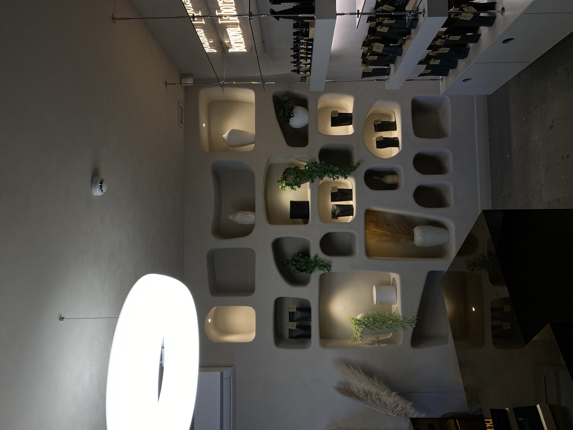 The image shows the interior of a space in Izhevsk, Russia, that features a wall display with irregularly shaped, recessed niches. Some niches contain decorative objects like vases, plants, and packaged goods. A tall, ring-shaped light fixture is visible against a light-colored wall. A counter or bar area with a dark, reflective surface is in the foreground, partially obscuring the lower right portion of the wall display. Overhead, there is a display of bottled products on shelves, and neon lighting fixtures. The environment is modern and stylish.