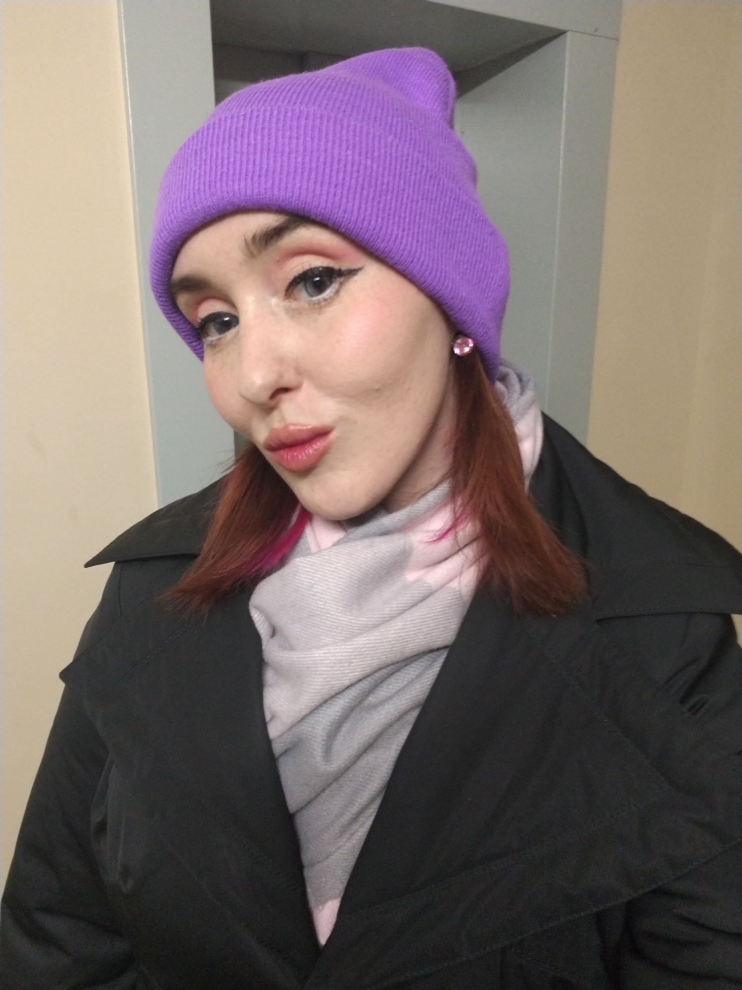 A person is wearing a purple beanie, pink eye shadow, a black coat, and a gray scarf. The location is not specified, so it is not possible to determine if it is Izhevsk, Russia.