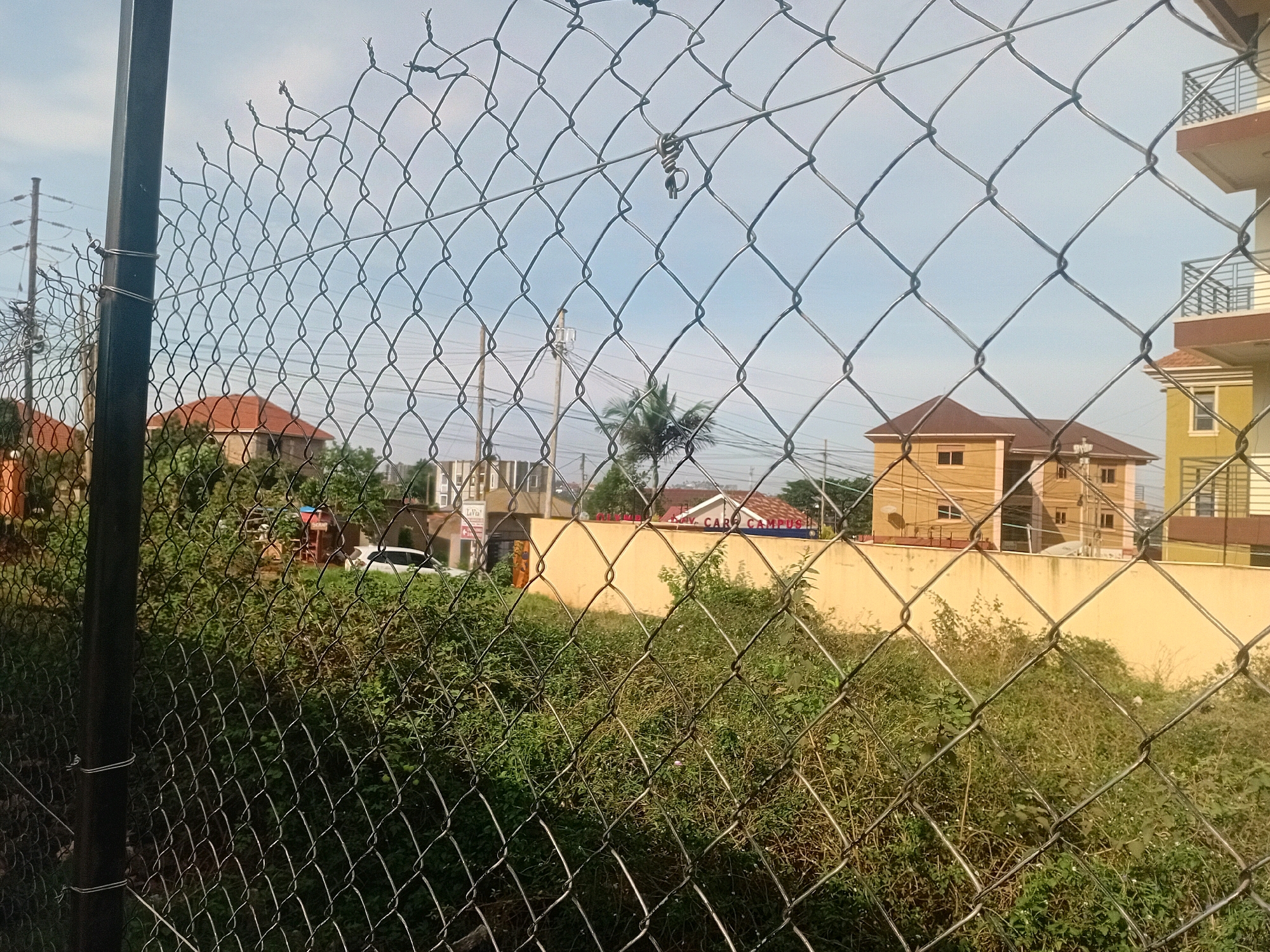 A chain-link fence obscures a view of residential buildings and overgrown vegetation in Kira, Uganda. The fence, made of twisted metal wires forming diamond shapes, is supported by a dark metal pole on the left. Beyond the fence, several buildings are visible, including a multi-story apartment complex with balconies and a smaller tan-colored house with a brown roof. The foreground is dominated by dense, green bushes and weeds. In the background, utility poles and wires are present, suggesting an urban or semi-urban setting. A white car is parked amidst the vegetation. The sky is overcast with a light blue hue.