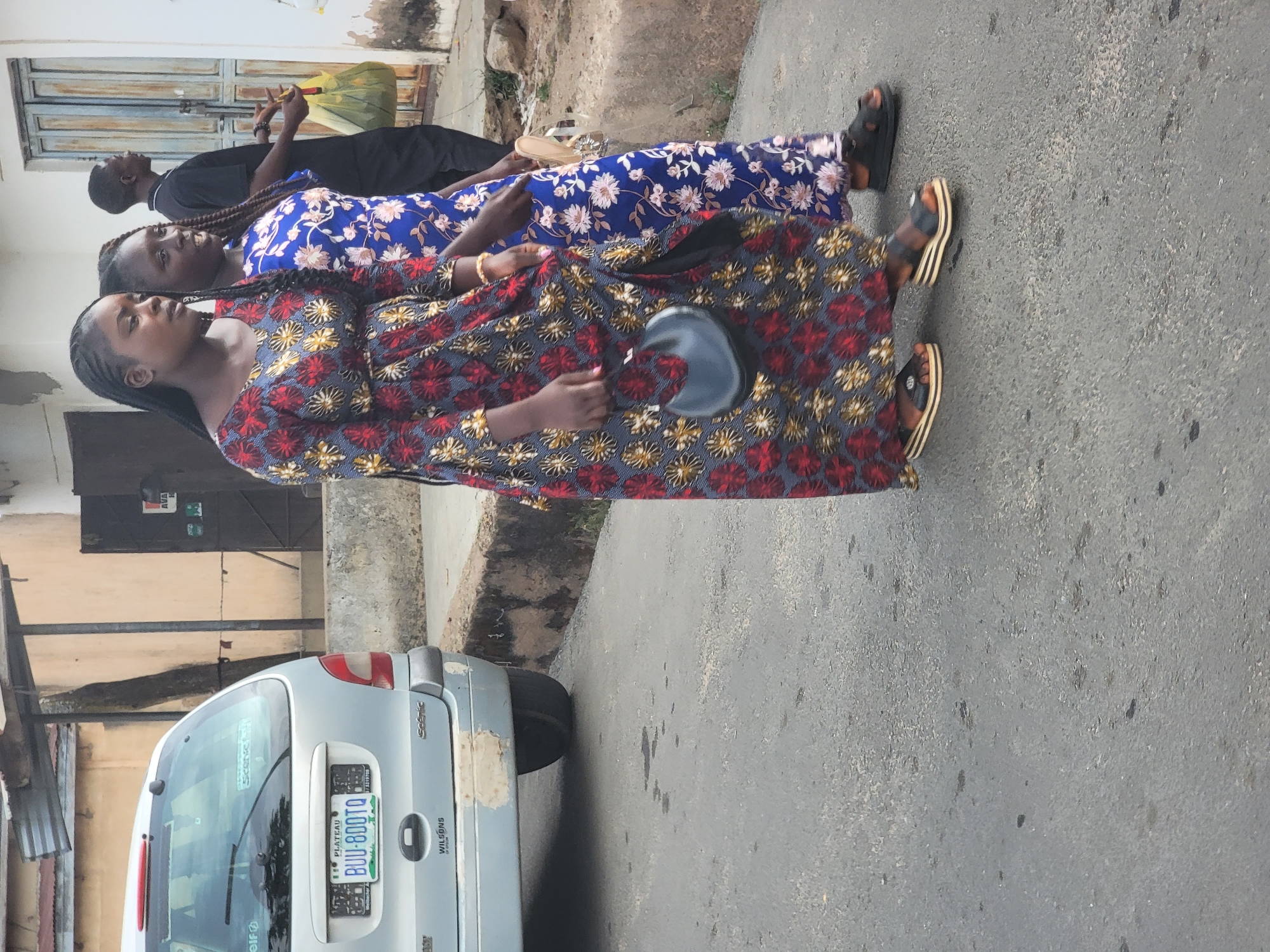 Three individuals are walking along a street. The first two are women wearing patterned dresses and sandals. The first woman is wearing a red and gold floral dress. The second woman is wearing a blue floral dress. Behind them is a man in black attire. A silver car is parked by the roadside. The license plate on the vehicle reads "BUU-800TQ," indicating registration in Plateau State, Nigeria. The setting is possibly residential, given the houses visible in the background. This image likely originates from the city of Jos, located in Plateau State, Nigeria.