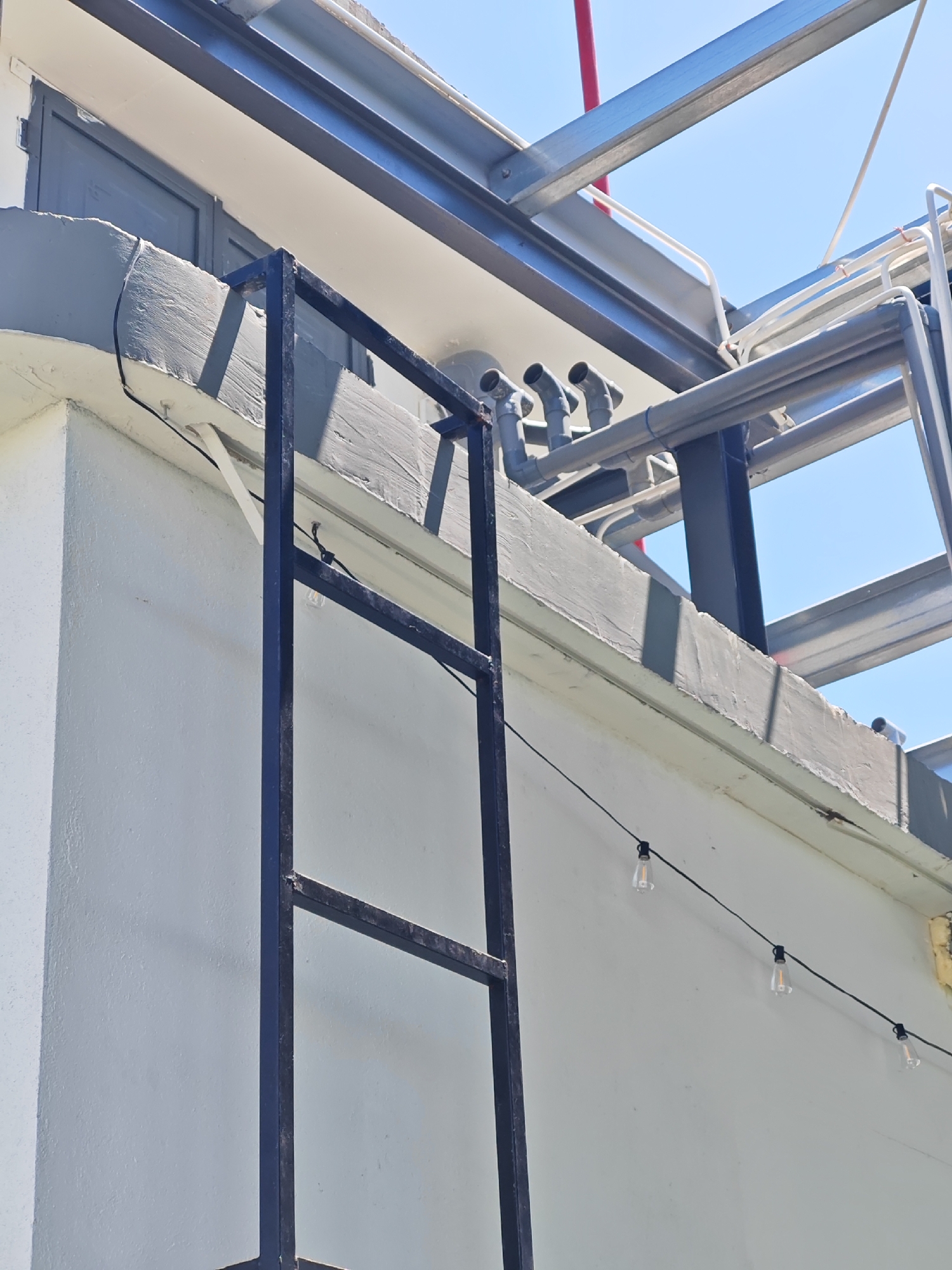 The image displays a close-up view of a white building's exterior in Đà Nẵng, Vietnam. A black metal ladder is partially visible, extending upwards against the wall. Above the ladder, there is a grey concrete ledge with some rough texture. A series of clear Edison-style string lights with black wires are strung across the building's facade. In the background, a complex structure of metal beams and pipes is visible against a clear blue sky. The lighting suggests it is daytime.