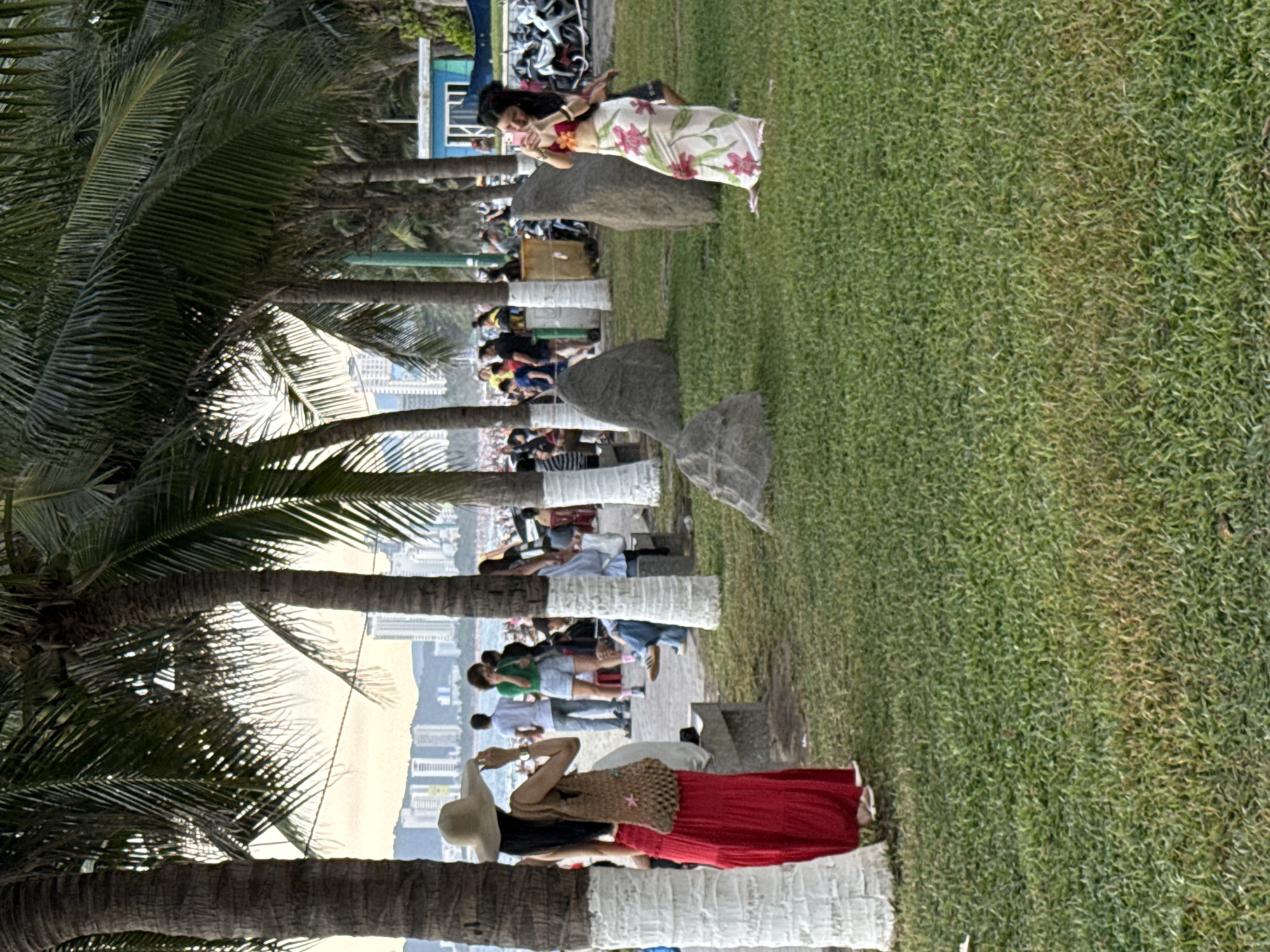The image depicts a park area adjacent to a beach in Nha Trang Ward, Vietnam. The foreground is dominated by a lush green lawn with scattered large, dark grey rocks and palm trees. The palm trees have their lower trunks painted white. In the middle ground, on a paved walkway, a woman in a red, flowing skirt and a wide-brimmed hat is facing away from the camera, holding a woven bag. Further along the walkway, other individuals are visible, some walking and others seated.
In the background, a beach is crowded with people, and beyond that, the ocean is visible. A cityscape with numerous tall buildings lines the horizon, indicating an urban setting. The sky is a soft, hazy orange, suggesting either sunrise or sunset. On the right side of the image, another woman is standing and taking a photo with her phone. She is wearing a floral-patterned skirt and a pink top. In the far right background, a row of motorcycles and a small blue building are discernible. The scene conveys a lively atmosphere at a popular coastal location.