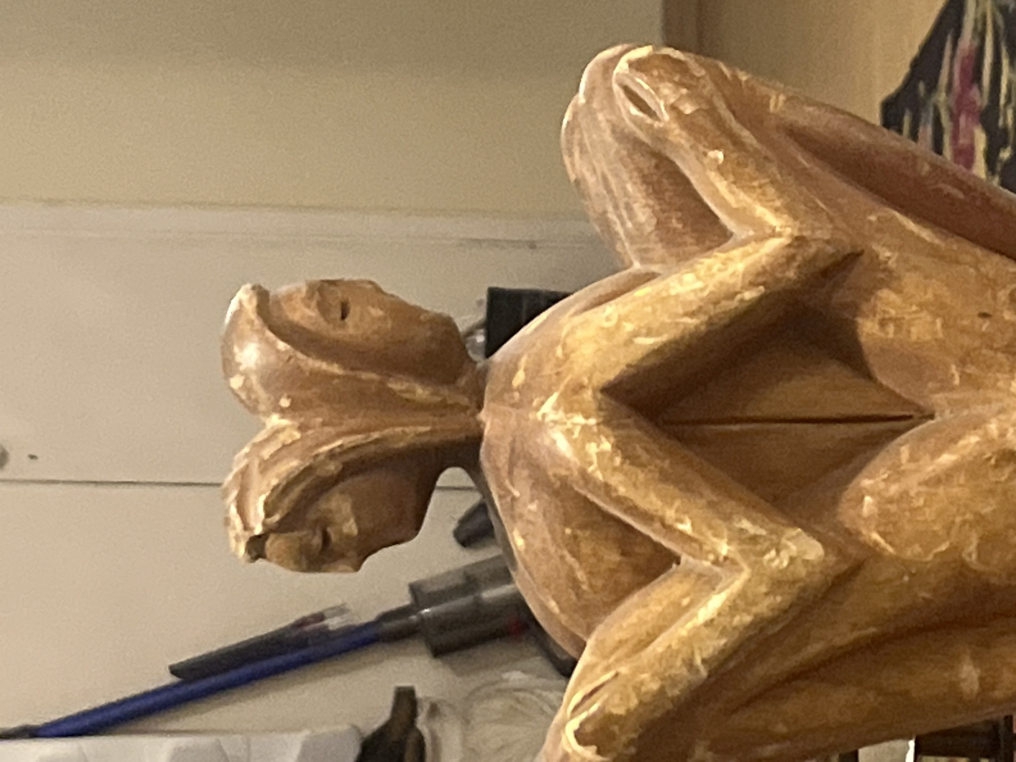 A close-up shot features a light brown, carved wooden sculpture. The sculpture seems to depict a figure with a head on the left side and stylized arms and legs forming a geometric shape on the right. The figure's head has carved features that suggest a face in profile. In the background, a white wall is visible, along with what appears to be a vacuum cleaner and other miscellaneous items on a surface. The image does not provide enough context to determine whether the location of this item is in Tel-Aviv, Israel, or any other city.