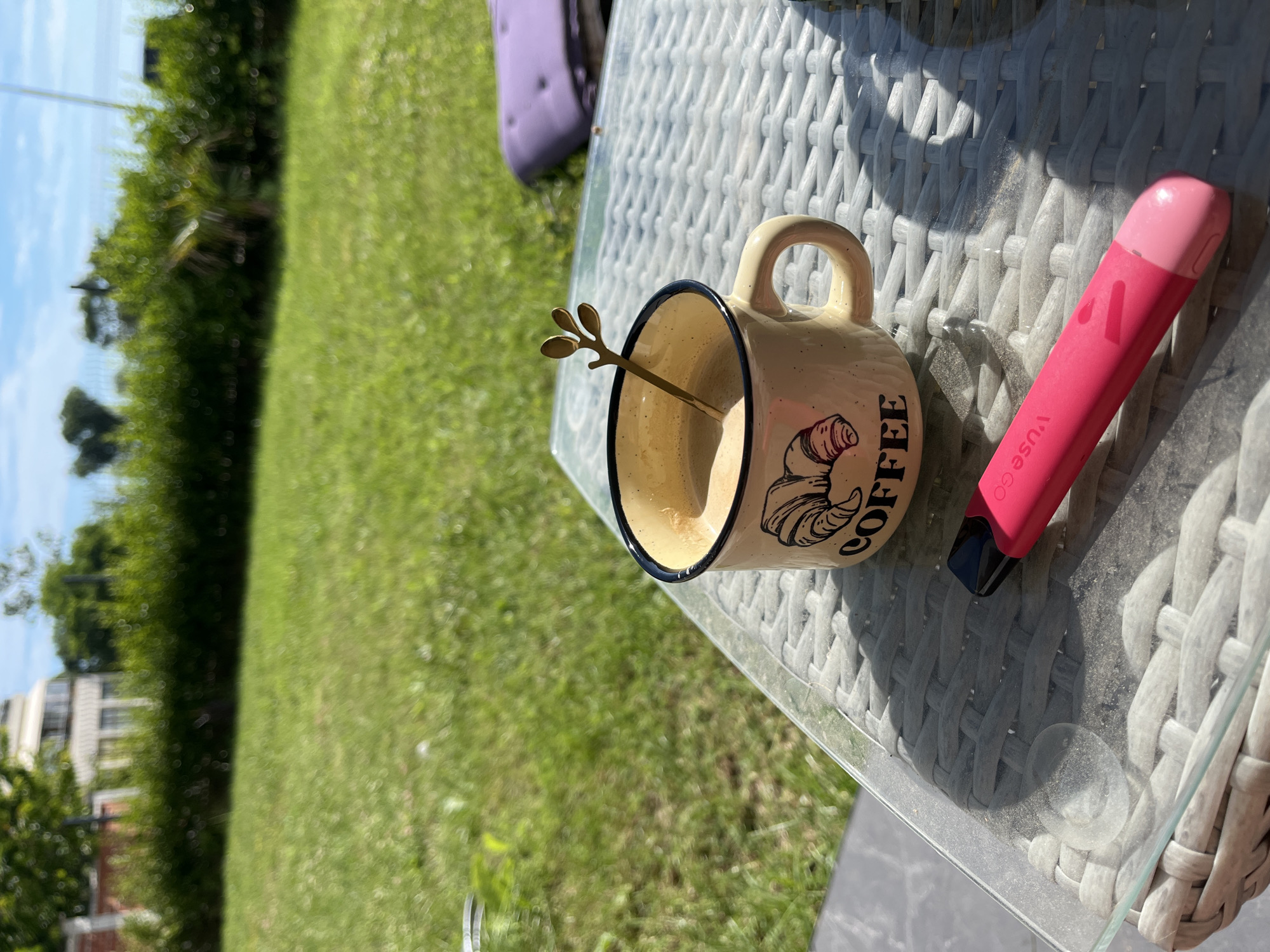 In Tsarevo, Bulgaria, on a sunny day, a close-up shot captures a coffee mug with a croissant illustration and the word "COFFEE" on its side, placed on a glass-topped wicker table. A decorative spoon with a leaf-like design is in the mug, stirring the coffee. A bright pink vape pen lies on the table in front of the mug. The background shows a lush green lawn, a hedge, and a building, suggesting an outdoor setting likely in a residential or public garden area.