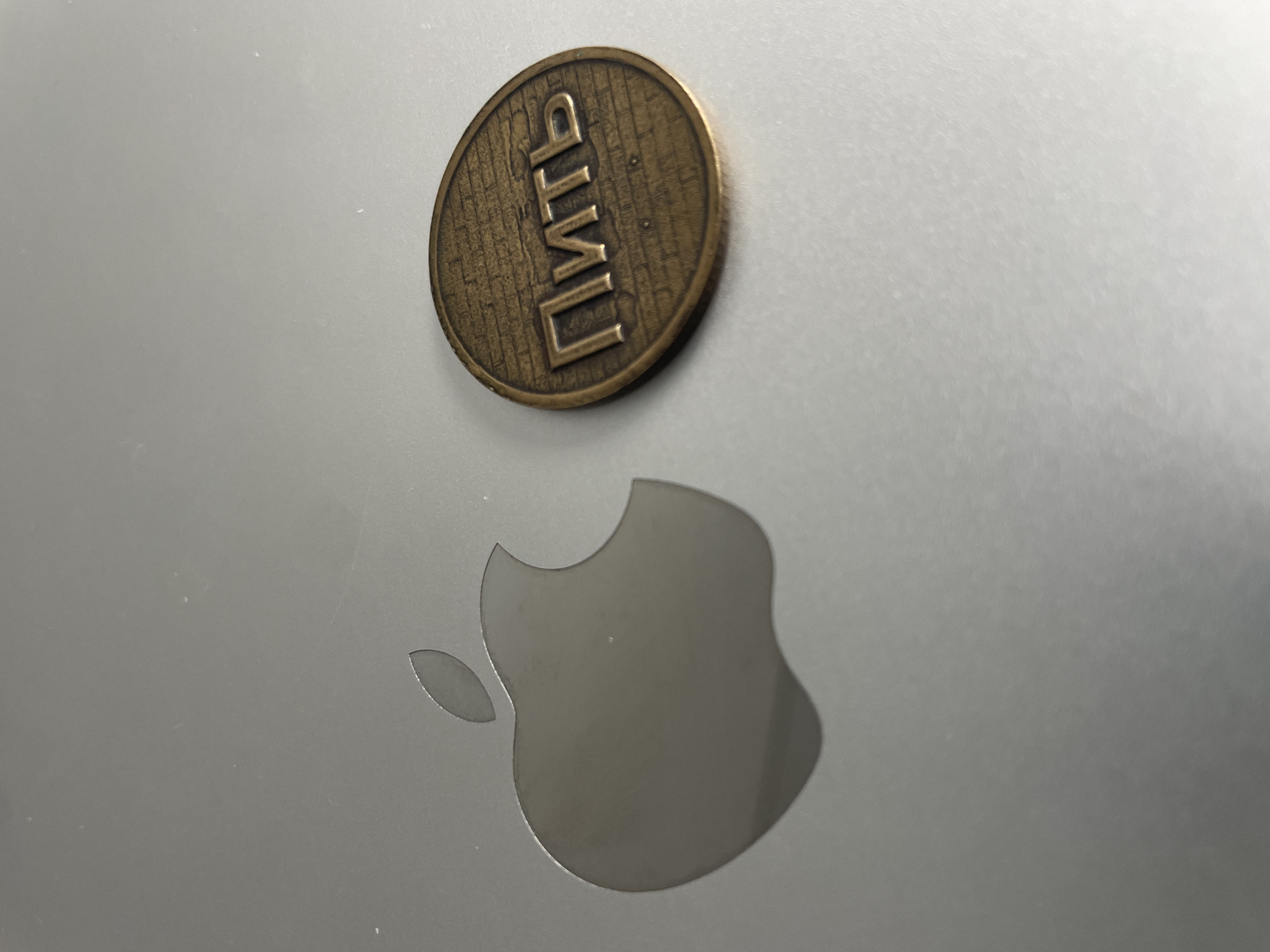 A close-up, eye-level shot shows a silver Apple logo etched into a metallic surface. To the right of the Apple logo, a round, bronze-colored coin with raised text is placed. The text on the coin, written in Cyrillic, appears to say "ПИТЬ", which translates to "to drink." The coin has a textured background that resembles bricks. The lighting suggests an indoor environment with a soft light source. There are no people visible in the image. The location is Unknown, Unknown.