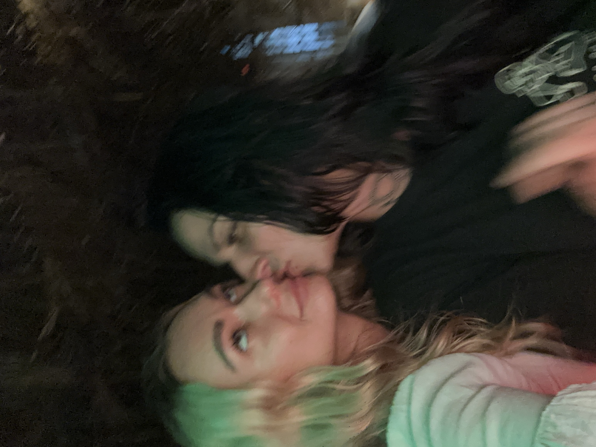 The image captures an affectionate moment between two individuals, likely young women, in a dimly lit setting at night in Mũi Né, Vietnam. The photograph is taken from a rotated perspective, suggesting the subjects are lying down or captured at an unconventional angle. One person, with long dark hair, is seen gently kissing the cheek of the other, who has long, wavy blonde or light brown hair. The person being kissed looks upwards, their face partially illuminated by a soft, greenish-blue light, creating a tender and intimate atmosphere.
The background is dark and heavily blurred, indicating low light conditions and possibly motion blur or a shallow depth of field. Faint, abstract reflections of artificial lights or neon signs are visible in the upper left area, suggesting an urban or entertainment environment. The individual with dark hair wears a dark top, with an indistinct, light-colored stylized graphic or design visible on their right shoulder or upper arm. The other person is wearing a light-colored, possibly white or light grey, textured garment on their arm. The overall blurriness of the image contributes to its candid and dreamlike quality, highlighting the emotional connection between the subjects. No explicit text is clearly legible within the image.