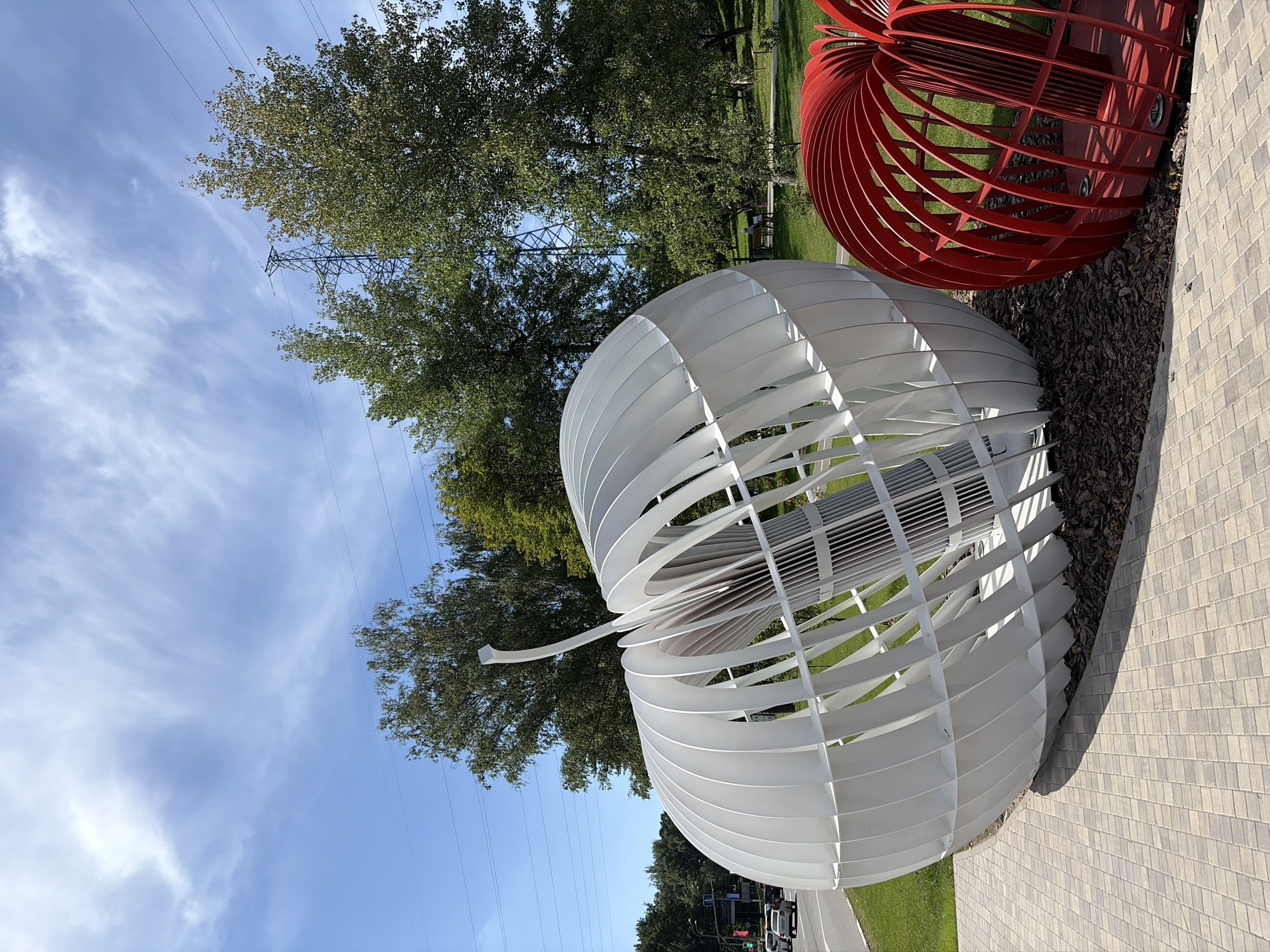 Two large, abstract sculptures resembling apples, one white and one red, are displayed outdoors in an urban park setting in Khimki, Russia. The white sculpture, dominating the foreground, is a deconstructed sphere with white, curved slats forming its outer shell. Inside, a cylindrical structure with vertical slats is visible. The red sculpture, partially visible to the right, is a similar deconstructed apple shape made of red curved slats. Both sculptures sit on a bed of mulch. The sculptures are placed on a paved walkway made of light-colored rectangular stones.
In the background, a street with cars driving on it is visible to the left, suggesting proximity to a road. Beyond the street, there are grassy areas and mature trees with green foliage. A power transmission tower and lines are visible above the trees, indicating the presence of infrastructure. The sky is blue with wispy white clouds. The overall scene suggests an outdoor public art installation in a park or green space within the city.