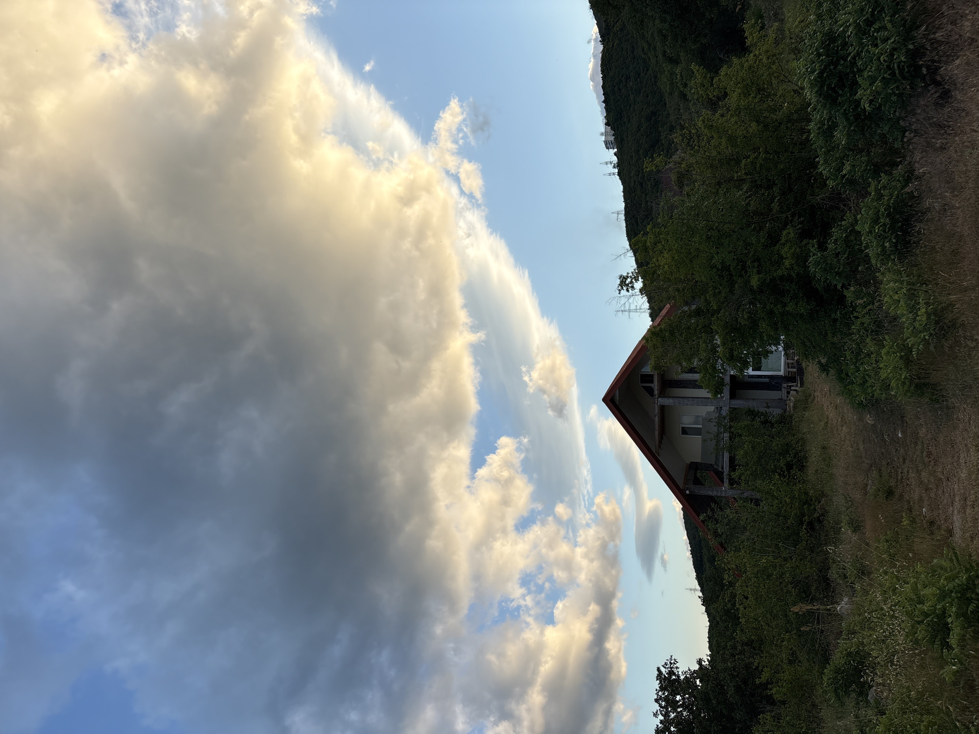 The image depicts an exterior view of a partially constructed house situated on a hillside. The house, made of concrete and with a red roof, is surrounded by dense green foliage and dry grass. In the background, a lush, green hillside is visible, dotted with trees and topped by structures that appear to be power lines or transmission towers. The sky above is a mix of blue and dramatic, large clouds, with some areas illuminated by the sun. The scene appears to be located in a rural or semi-rural area, possibly in Malyi Mayak, Ukraine, given the presence of these specific environmental and architectural features. There are no people or animals visible in the image, and the overall atmosphere suggests a natural, somewhat undeveloped landscape with ongoing construction.