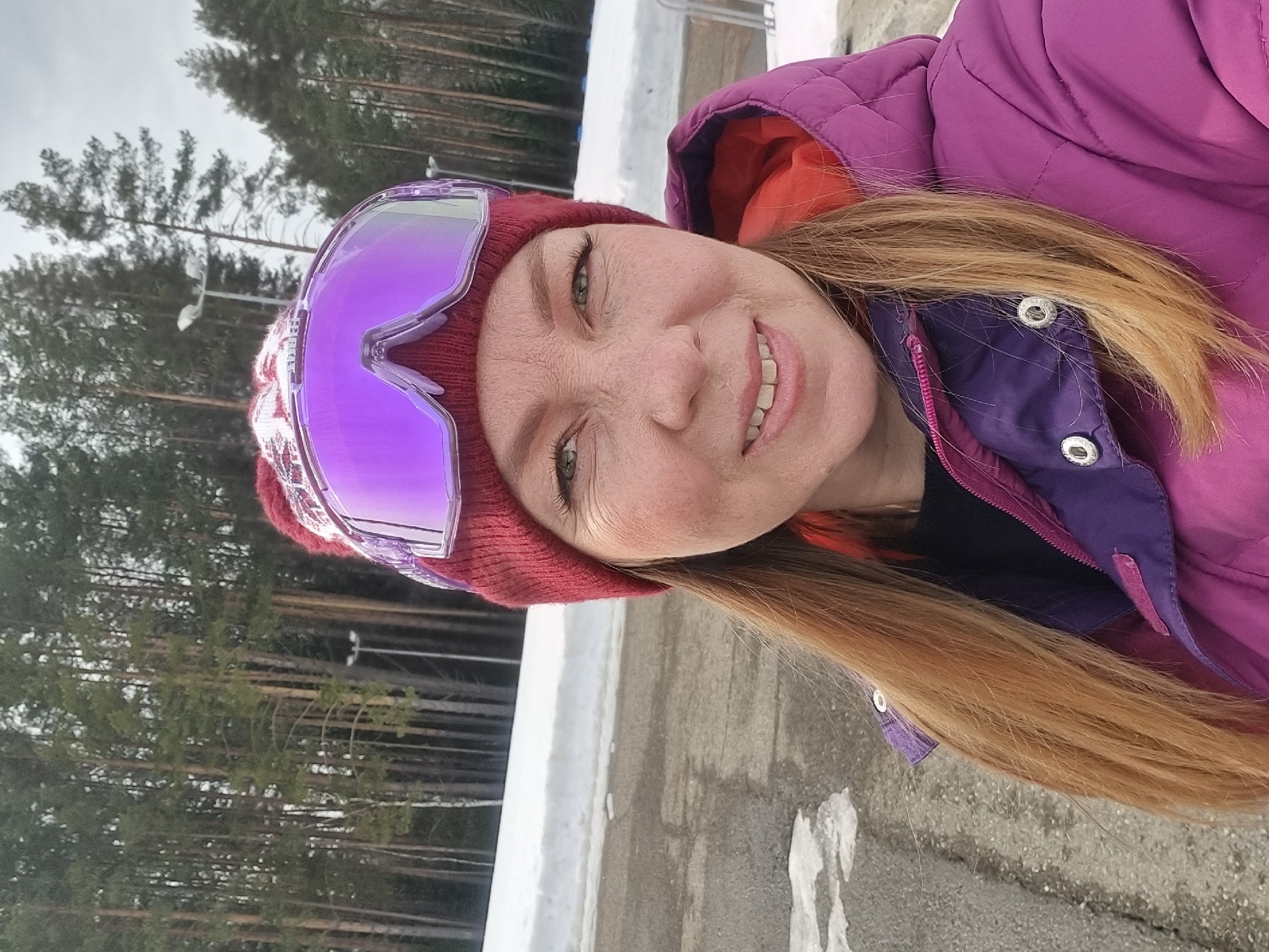 A female individual is depicted in a close-up, selfie-style outdoor portrait within what appears to be a recreational area in Прикамский, Russia. She has light-colored hair, green eyes, and visible freckles, and is looking directly at the camera with a slight smile, revealing her teeth. She wears a dark red knitted beanie with a white patterned design on the front, adorned with reflective purple-lensed ski goggles pushed up onto her forehead. Her attire also includes a quilted magenta or purple winter jacket with an orange inner lining visible around the collar, worn over a dark garment. The jacket features silver-colored snap buttons on a purple flap near the shoulder. The background consists of a dark, partially snow-covered paved surface in the midground, with a distinct snowdrift bordering the right side of the frame. Beyond the snowdrift and paved area, a dense forest of tall, slender evergreen trees is visible under an overcast sky. Several tall poles with streetlights or floodlights are interspersed within the treeline. The overall environment suggests a winter or early spring outdoor setting, likely a ski resort or forested park.