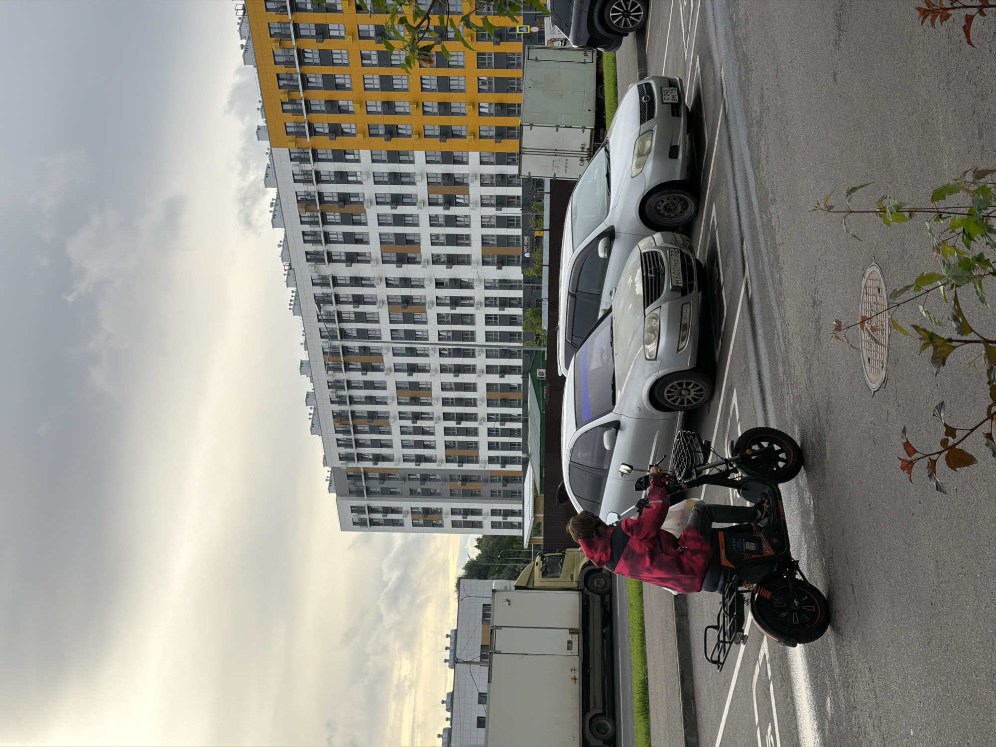 A person rides an electric scooter down a street in Kazan, Russia. The person is wearing a red and black hooded jacket and dark pants. They are carrying a white bag. The street is asphalt with white lane markings. In the background, there is a large modern apartment building with a yellow and white facade, and several cars parked in a lot. To the left of the apartment building, there is a white delivery truck. The sky is overcast.
