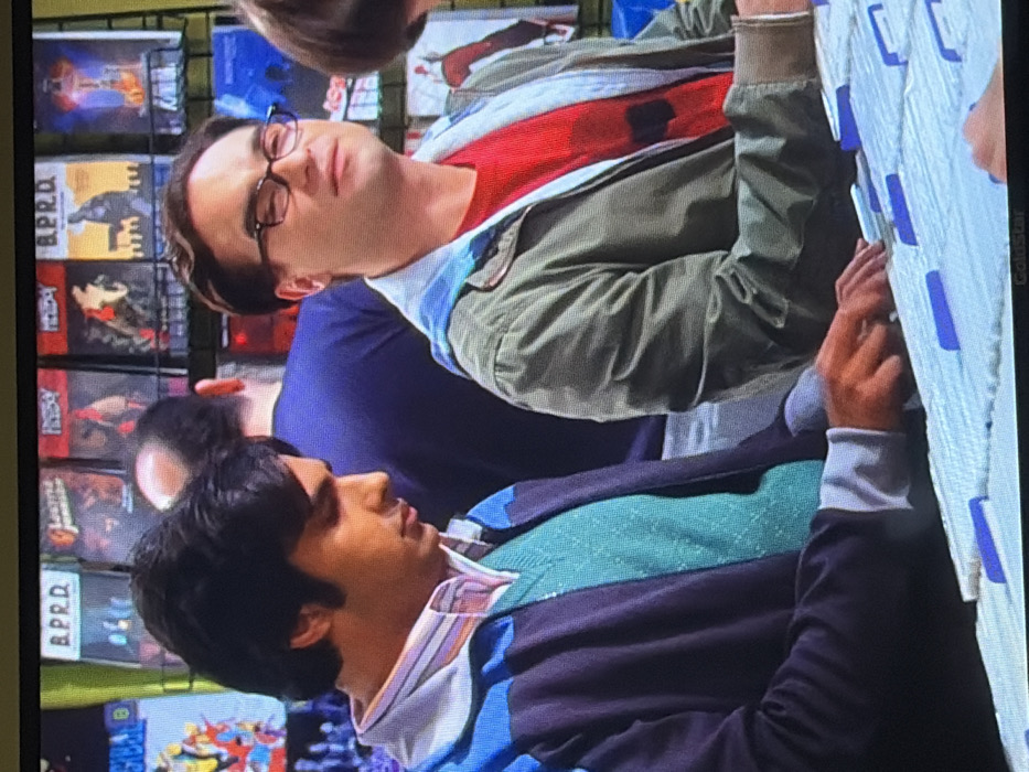 Two men stand side-by-side, facing left. The man at the top wears glasses, a red shirt, and a green jacket. The man at the bottom wears a collared shirt, a turquoise sweater, and a blue jacket. Behind them are shelves filled with various items, including what appear to be comic books with visible titles like "B.P.R.D." and "History." The men appear to be looking at items hanging on a rack with blue labels.