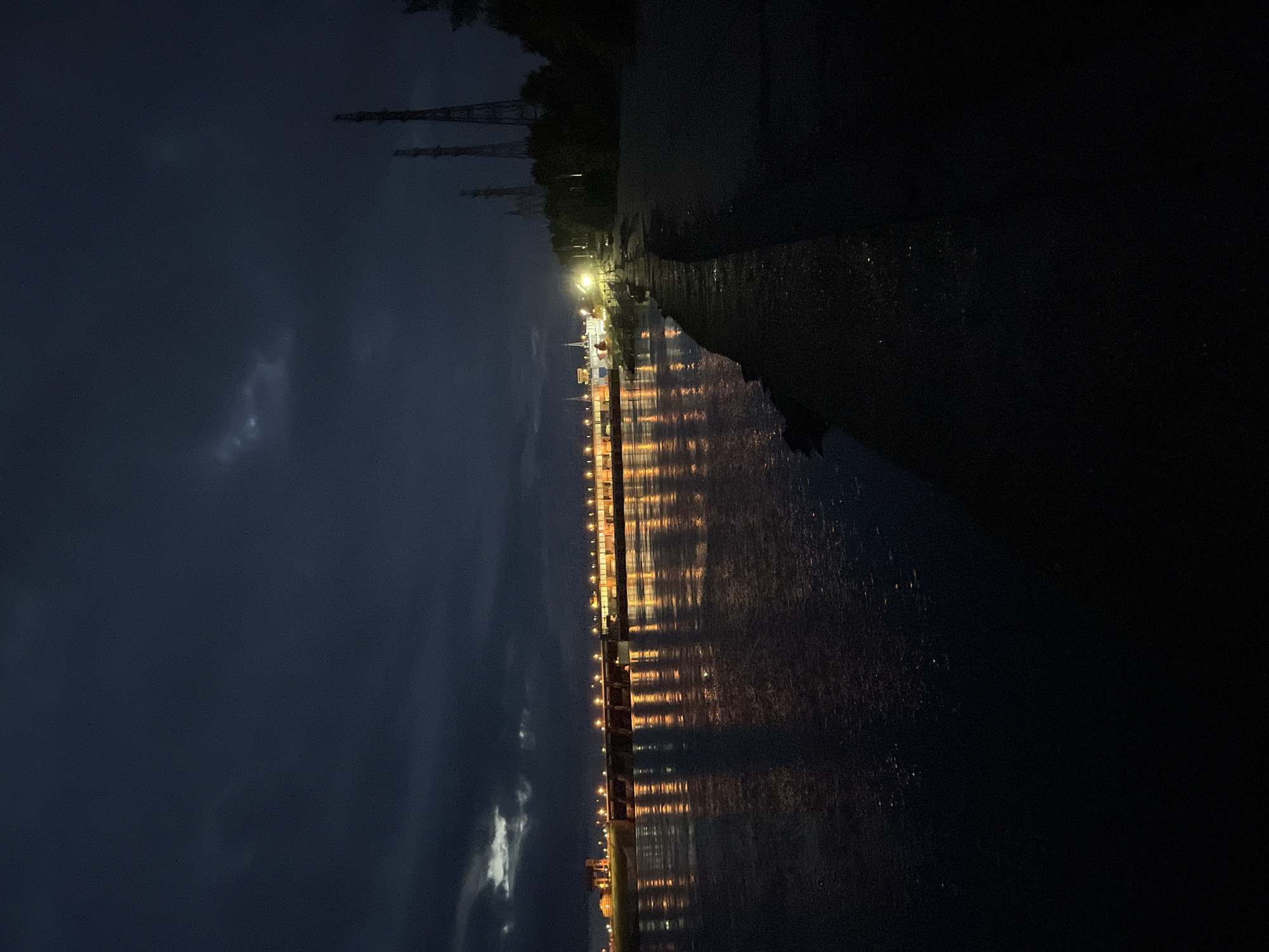 The image captures a nighttime view of the Votkinsk Hydroelectric Station on the Kama River in Chaikovsky, Russia. The sky is dark and heavily overcast, with faint lighter cloud formations visible in the upper left quadrant. A wide river dominates the lower and middle sections of the frame, its surface reflecting numerous warm-toned artificial lights from the dam. The dam structure, spanning the river horizontally in the mid-ground, is brightly illuminated, revealing its gate sections and associated buildings. On the right side, a dark, textured embankment or shoreline extends from the foreground towards the distant dam. Several tall, slender industrial towers are partially visible behind the right shoreline, silhouetted against the dark sky, with dense foliage obscuring their lower sections. No people, vehicles, or specific activities are identifiable within the scene.