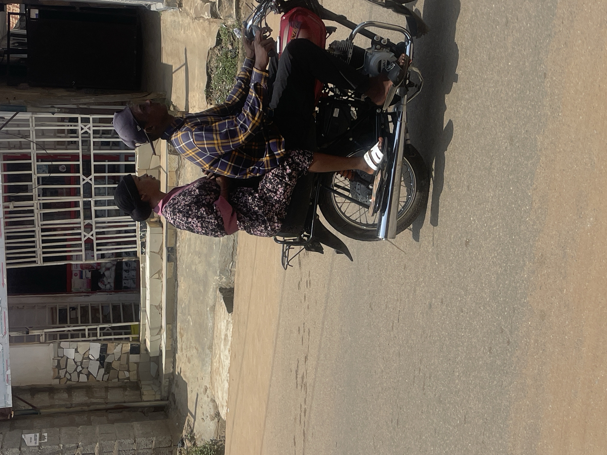In Abuja, Nigeria, a young man and woman are riding on a red motorcycle. The man, wearing a blue and yellow plaid shirt and a dark blue baseball cap, is driving. The woman, in a floral print dress and a black headscarf, is a passenger. They are traveling on a paved road, with buildings and shops visible in the background. The scene appears to be a typical day in a bustling urban environment.