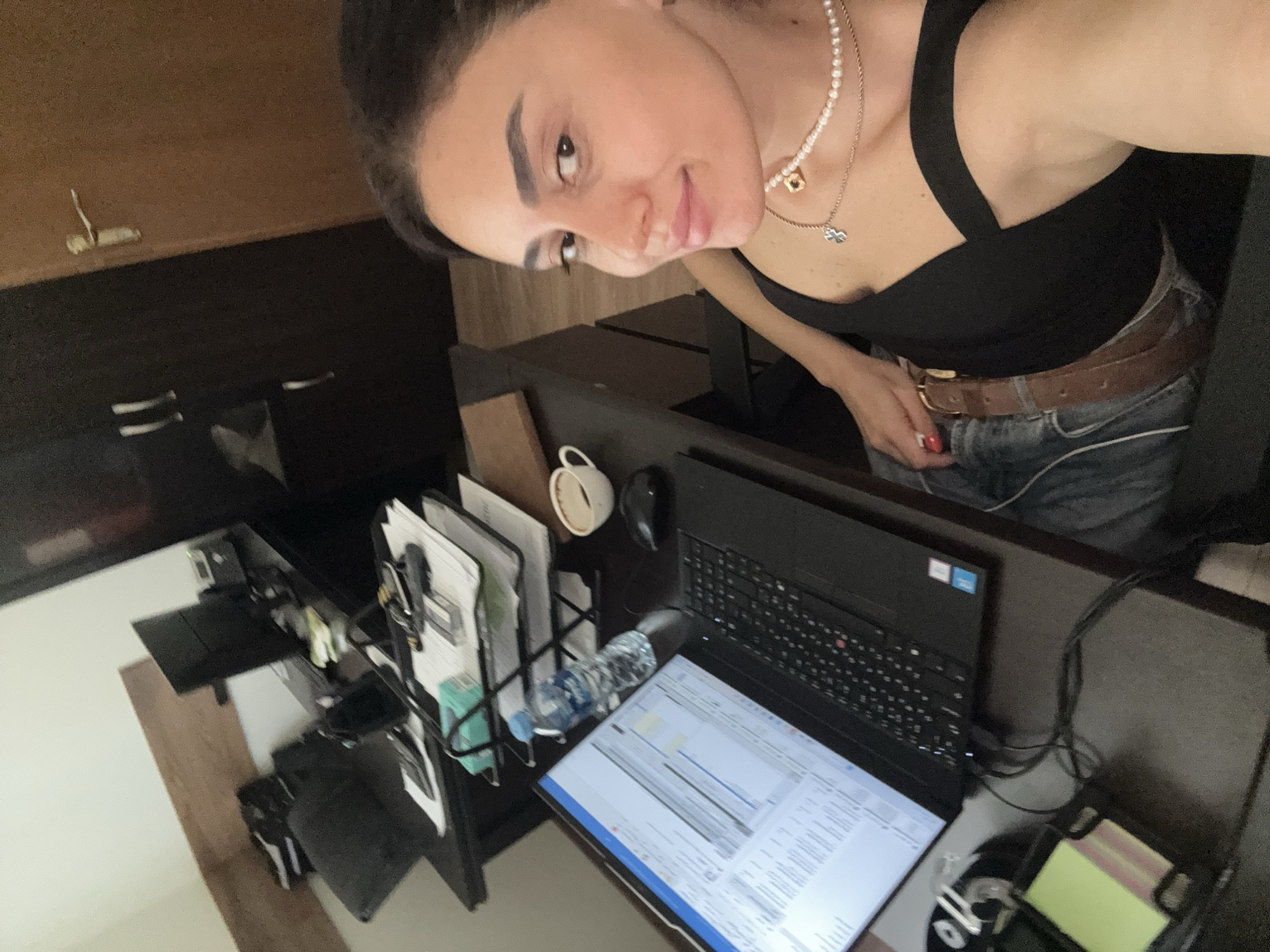 A woman is sitting at a desk in what appears to be an office or home office environment in Tbilisi, Georgia. She is looking at the camera, smiling. In front of her is an open laptop with a spreadsheet displayed on the screen, a bottle of water, a white cup with coffee, and a computer mouse. Behind the laptop, there are stacks of papers in a black wire organizer, and further back, there is a computer monitor and a telephone. The desk appears to be dark wood. To the left of the desk is a black office chair. In the background, there is a dark wooden wardrobe with two doors, and a wooden door with a gold handle. The walls are painted white and there is a wooden plank mounted on the wall above the desk. The woman is wearing a black top, jeans, a brown belt, and two necklaces.
