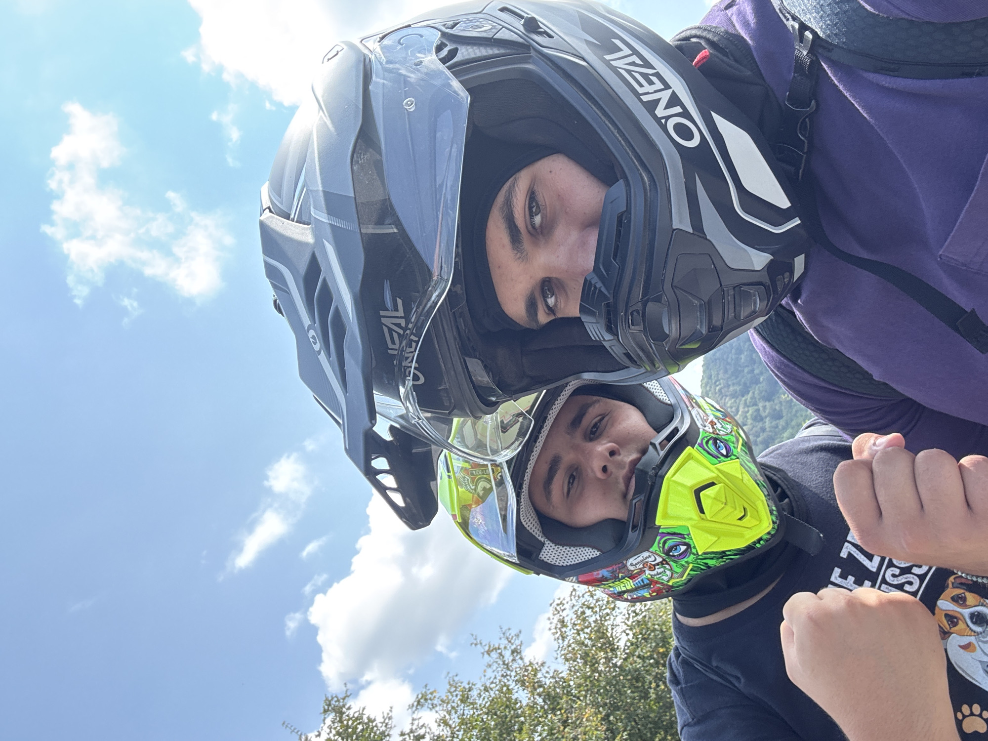 Two young men wearing motocross helmets are taking a selfie outdoors. The man on the left has a brightly colored helmet with green, yellow, and various cartoonish graphics. He is wearing a black t-shirt with a graphic design featuring dog faces and text. His right fist is clenched and held towards the camera. The man on the right wears a predominantly black and gray helmet with the "O'NEAL" logo visible. He is wearing a purple long-sleeved shirt and a black backpack. His left fist is also clenched and raised. Both men are looking towards the camera. The background consists of a blue sky with scattered white clouds, green foliage from trees on the left, and a blurred hillside in the distance. The image appears to have been taken on a sunny day. The location is Chelopek, Bulgaria, a village in the western part of the country.