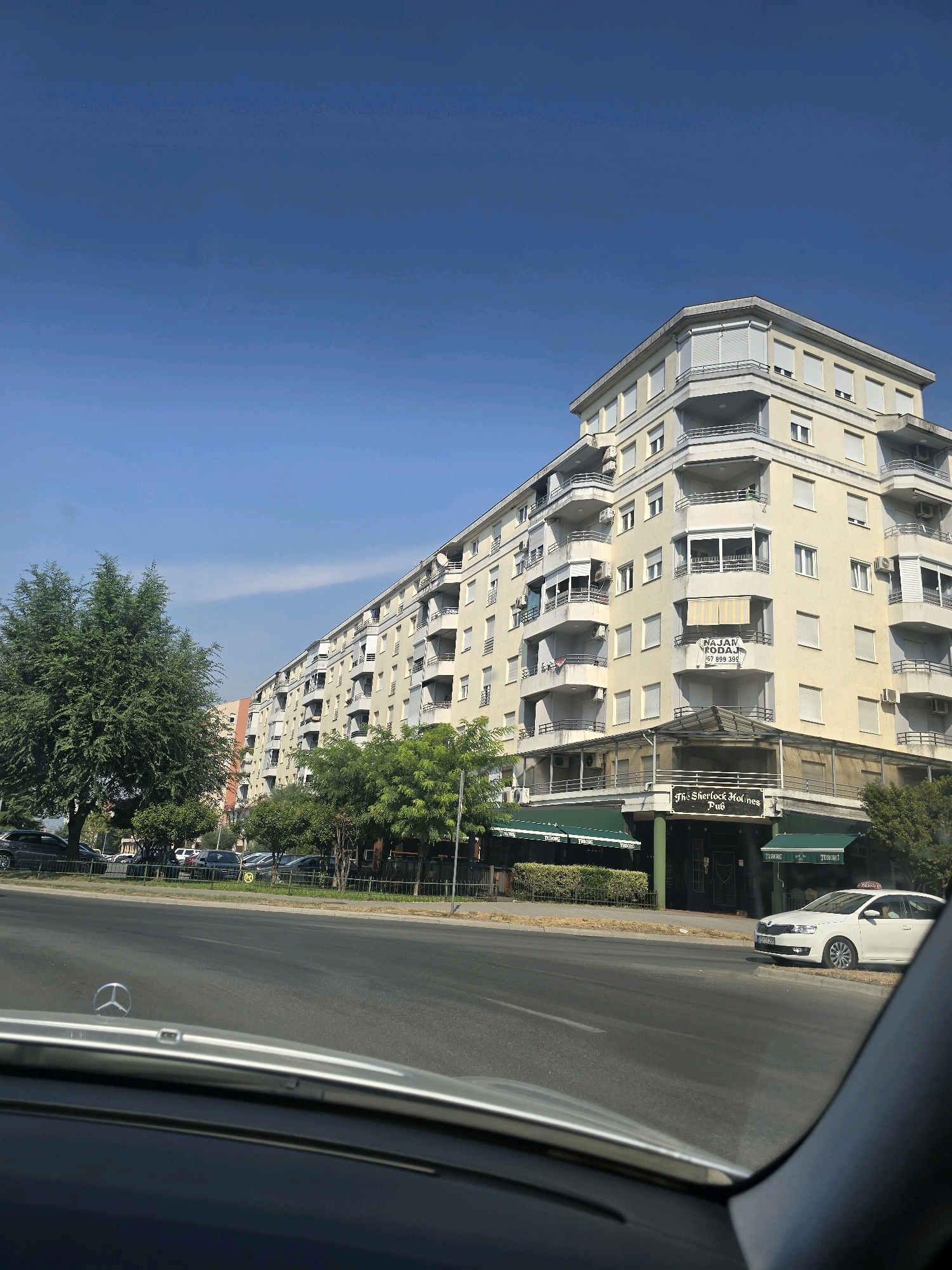 The image shows a view from inside a car looking out at a long, cream-colored apartment building in Podgorica, Montenegro. The building has multiple balconies with railings and closed shutters on many of the windows. There are trees and bushes in front of the building and along the street. A street sign is visible on the building that reads "The Sherlock Holmes Pub". A white car with a taxi sign on its roof is parked on the side of the road. The sky is clear and blue.