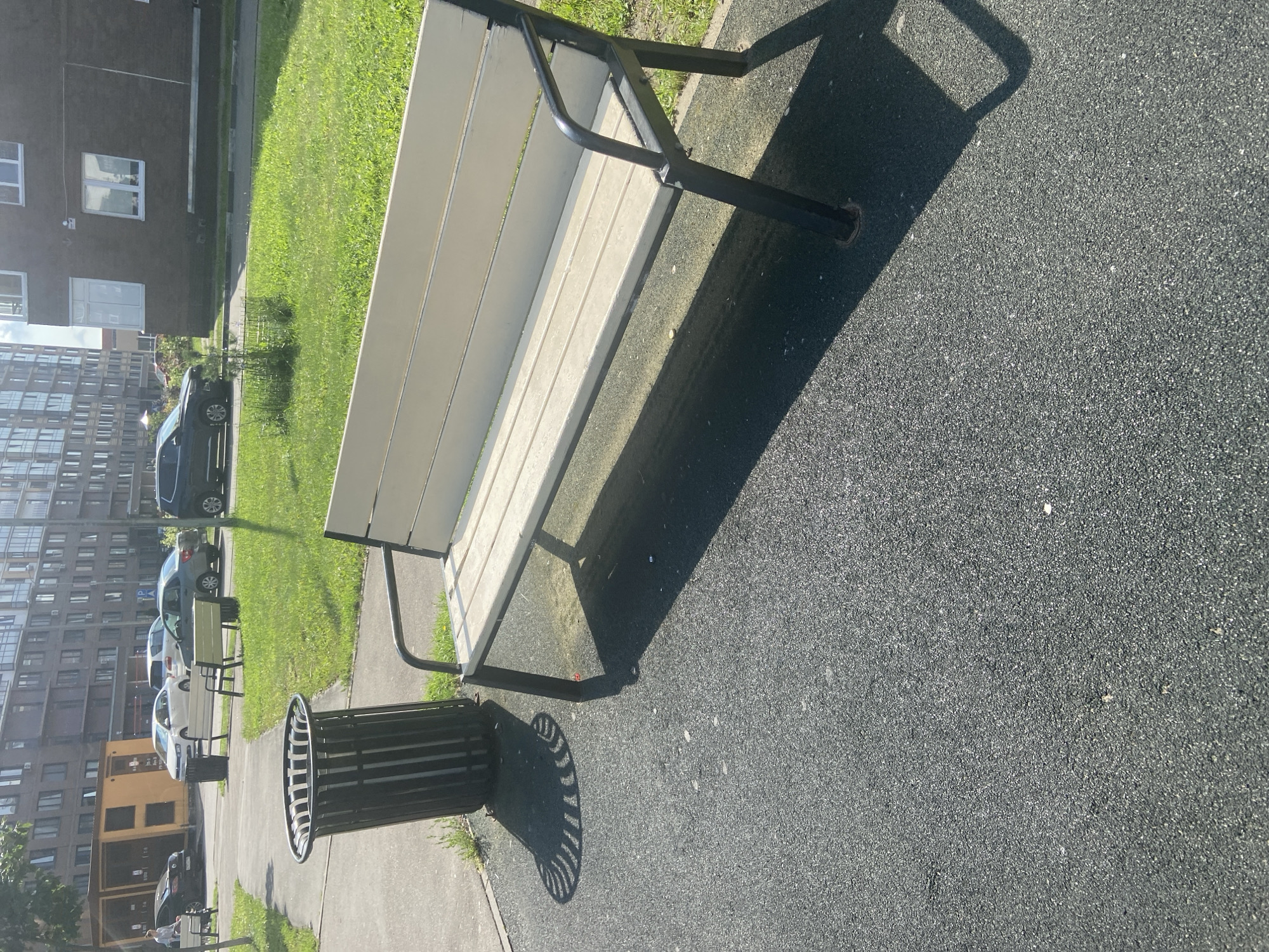 A street scene in the city of Balashikha, Russia, features a park bench positioned on a dark asphalt surface in the foreground. The bench is made of light-colored wooden slats and has black metal legs and armrests. To the left of the bench, a black cylindrical trash can with a slatted design stands on the asphalt, casting a patterned shadow. In the background, a grassy area with scattered trees is visible, beyond which are apartment buildings and parked cars. A white car is parked in front of a yellow modular building on the left. The sky is bright and appears to be a clear day.