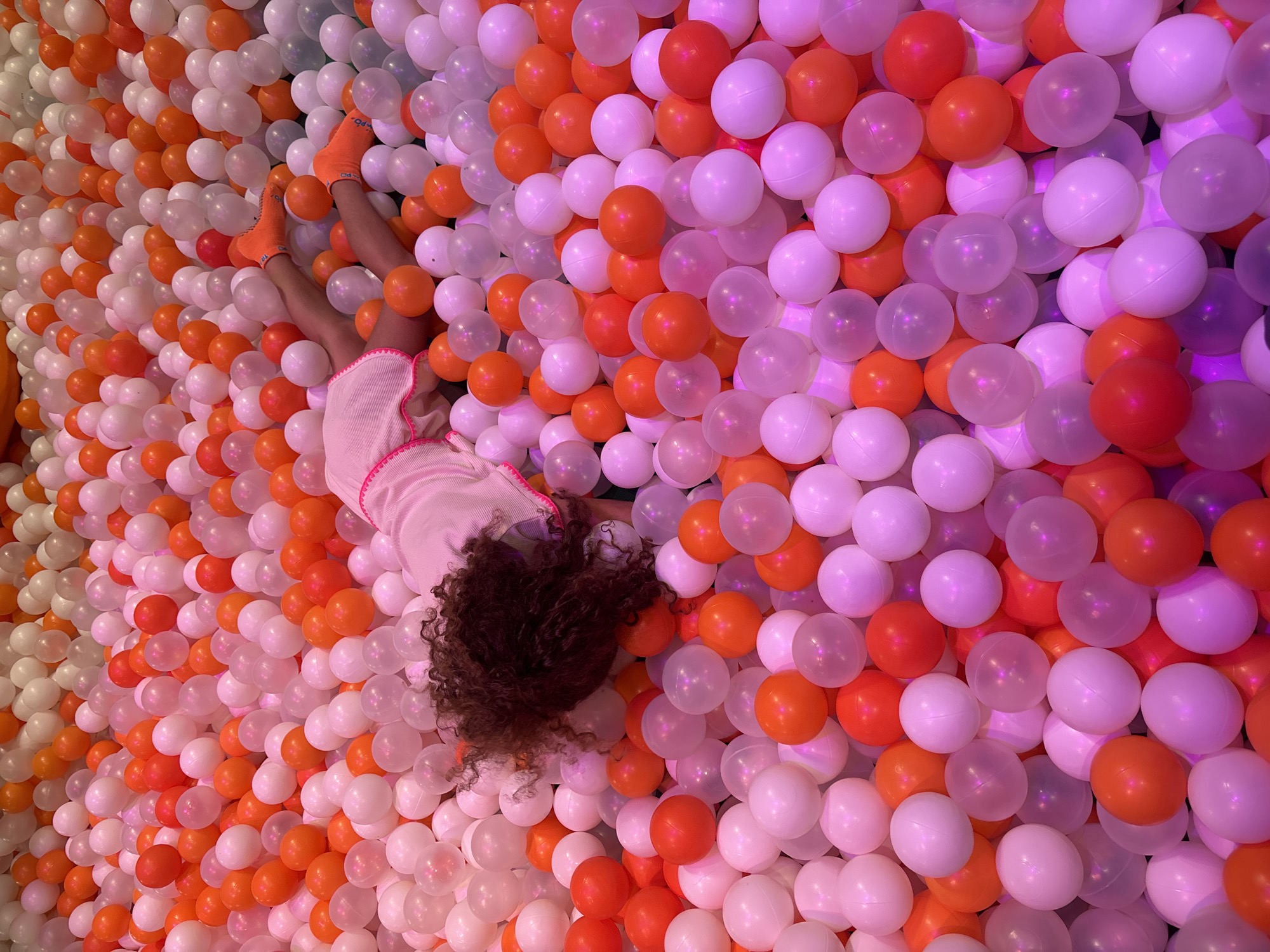 The image shows a young child lying on their back in a ball pit filled with numerous small, round balls of varying colors, primarily white, orange, and pink. The child is wearing a light-colored dress with pink trim and orange socks. Their face is not visible as they are looking down into the ball pit. The ball pit appears to be an indoor play area, possibly at a family entertainment center or theme park. The lighting is soft and appears to have a pinkish-purple hue, contributing to the playful atmosphere. There is no information in the image to suggest a specific location in Dubai, United Arab Emirates.