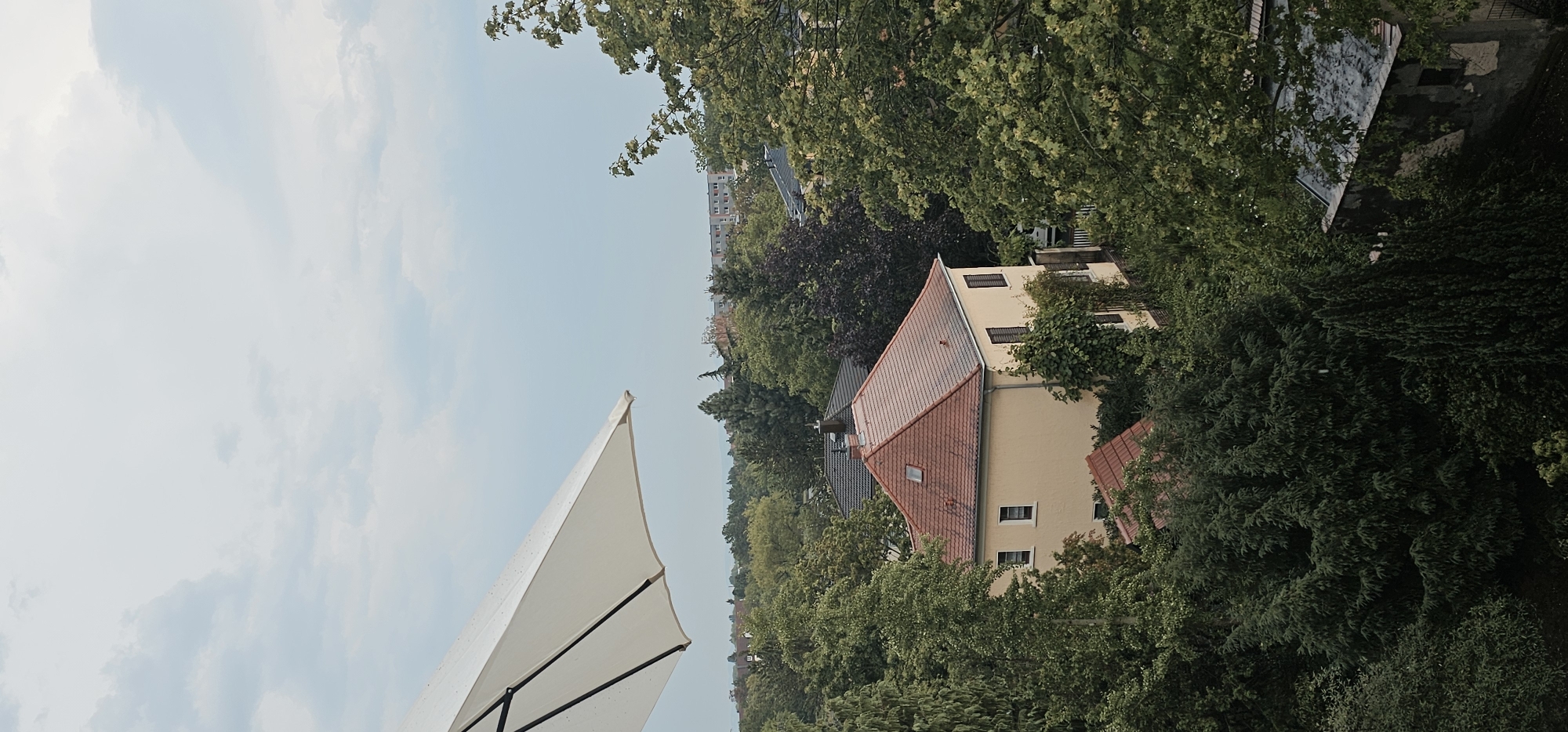The image shows a view from a balcony or elevated position overlooking a residential area with dense greenery. A large white umbrella is partially visible in the upper left corner, angled downwards. Below the umbrella, a light yellow house with a red tile roof stands out amongst a variety of trees and other buildings. The house has several windows and shutters. In the background, more buildings and a hazy sky with clouds are visible. The scene appears to be a quiet, suburban landscape. The image does not contain any people or specific actions.