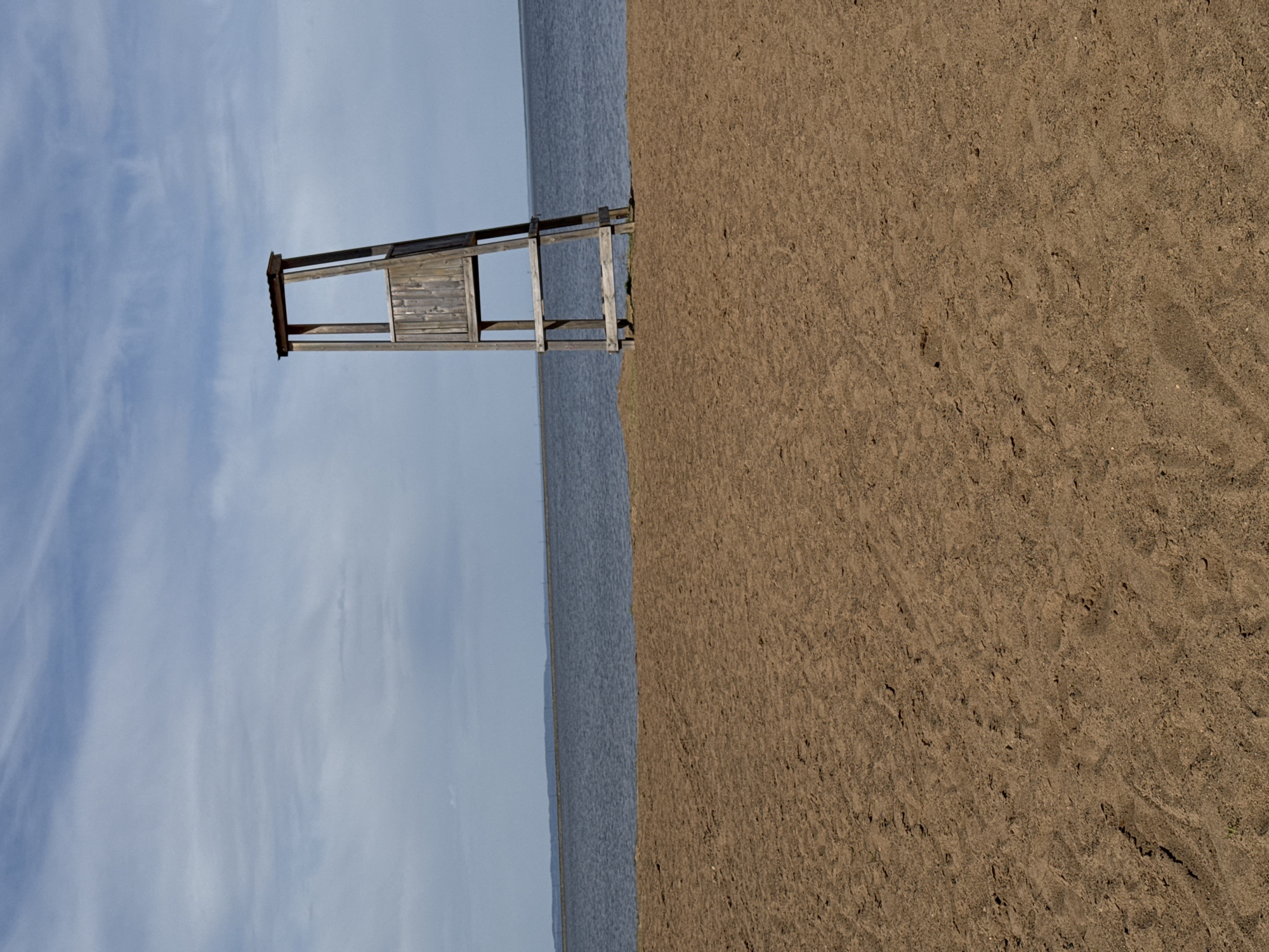 A photograph taken at the beach in Chaikovsky, Russia, presents an outdoor scene featuring sand, water, and sky. A wooden lifeguard tower is the central element. The tower straddles the edge of the water, with its base seemingly rooted in the sand. The water, with a slightly rippled surface, stretches towards the horizon. The sky is visible in the background, exhibiting light clouds. The sand texture is coarse, and its coloration is a muted brown. The image is rotated 90 degrees counter-clockwise.