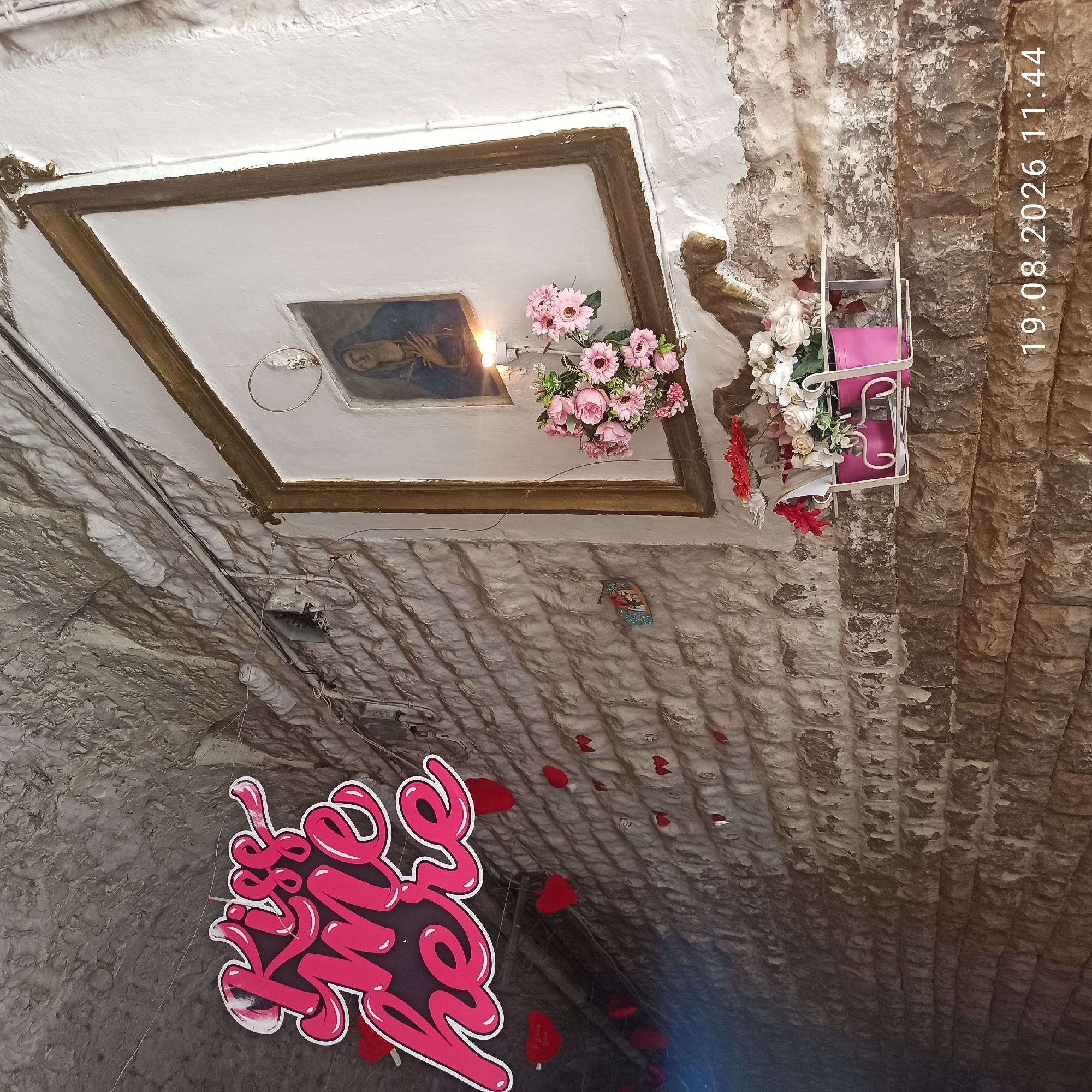The image depicts a narrow, arched alleyway in Ruvo di Puglia, Italy, characterized by rough-hewn stone walls and a textured ceiling. A large, pink, neon-style sign with the words "Kiss me here" is prominently displayed on the left side of the frame, suspended by a thin wire. Below the sign, numerous small red heart decorations are strung along a wire, with one larger heart visible on the left.
On the right side of the alley, a framed picture of the Virgin Mary is hung on a white plastered wall, with a candle burning below it, casting a warm glow. A bouquet of pink artificial flowers is placed on a shelf beneath the framed image. Further down the wall, a smaller arrangement of red and white artificial flowers in a pink pot is also visible. Electrical conduits and wiring are visible along the textured ceiling and walls, suggesting the presence of modern infrastructure within the ancient setting. The lower right corner of the image displays a timestamp: "19.08.2026 11:44". No people are present in the scene.