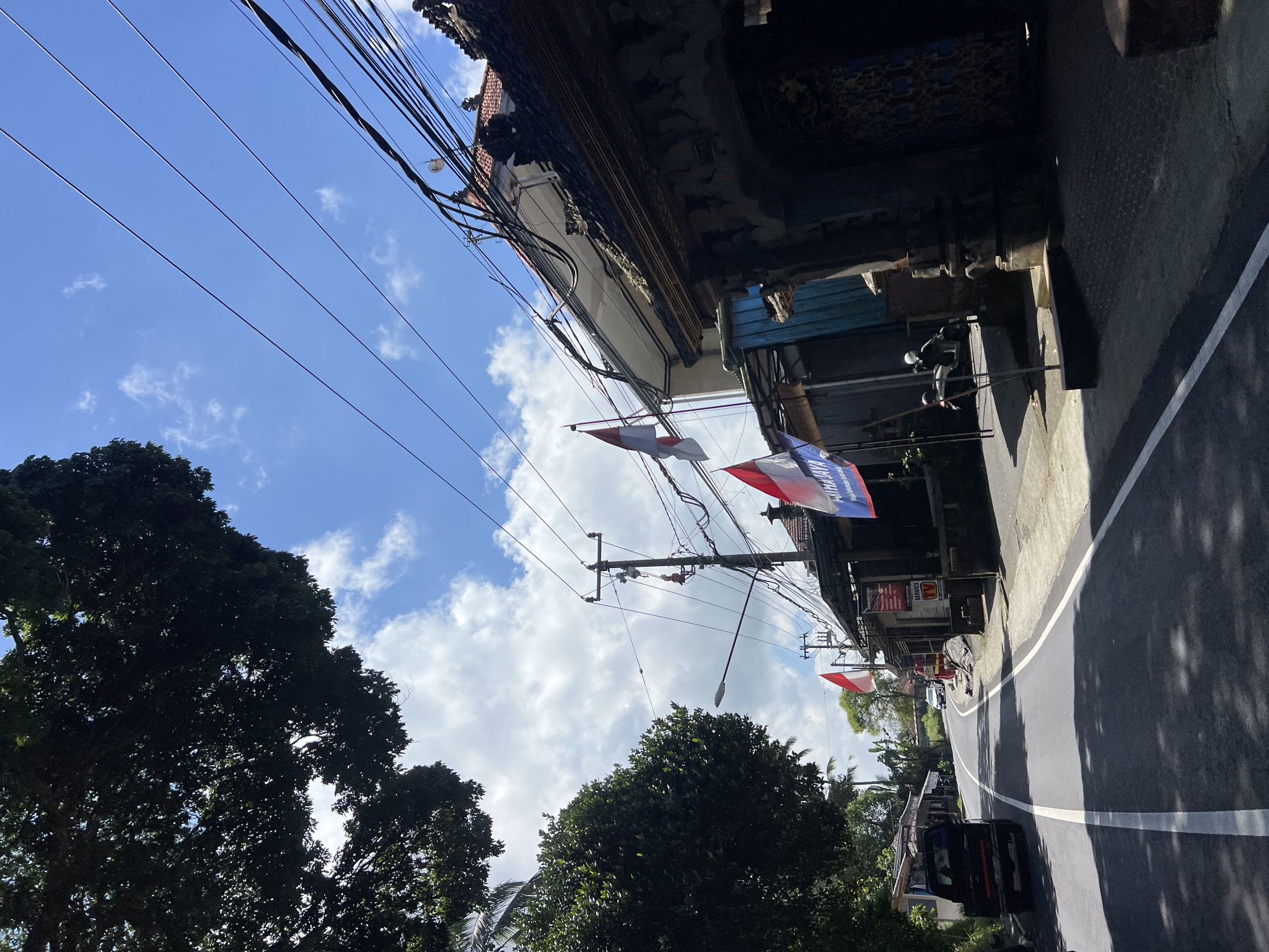 In Payangan, Indonesia, a quiet, paved road winds through a sparsely populated area lined with traditional Indonesian architecture and lush vegetation. The sky above is a vibrant blue, dotted with fluffy white clouds, and framed by the dark canopy of a large tree on the left. Power lines crisscross the sky, supported by utility poles.
On the right side of the road, several buildings display the red and white Indonesian flag, indicating a festive occasion. The structures feature intricate carvings and decorative elements characteristic of Balinese design. Some buildings appear to be shops, with one displaying a sign for "SERVICE TV." A motorcycle is parked in front of one of the establishments.
On the left side of the road, a black pickup truck is parked. Further down the road, another car is visible in the distance. The road itself is marked with white lines and is shaded in parts by the trees. The overall scene suggests a tranquil day in a Balinese village.