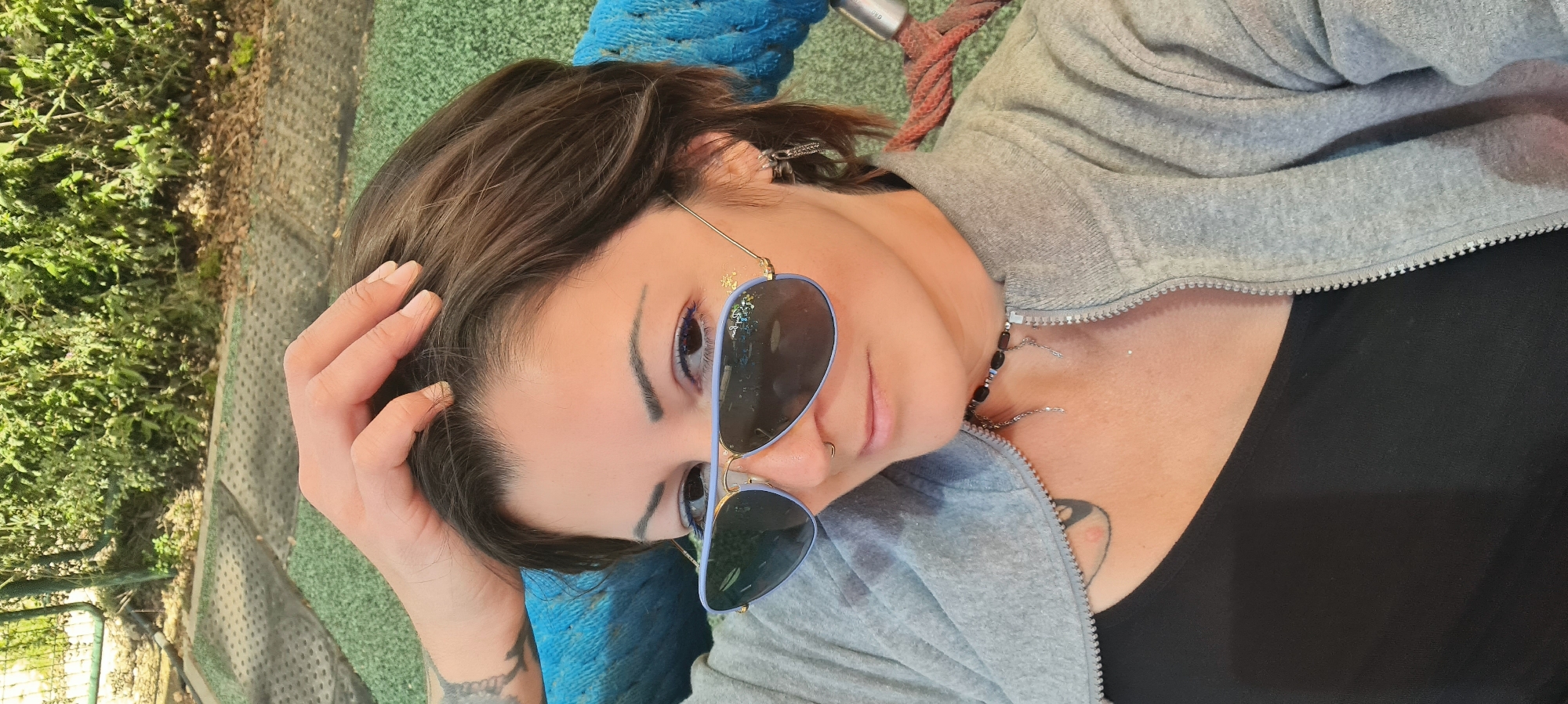 A woman lies on a blue rope structure on green synthetic turf in an outdoor area. She has light skin, brown hair, and is wearing sunglasses, a grey zip-up hoodie, and a black top. She has a hand on her head and is looking towards the camera. Behind her is a grassy area with some bushes.