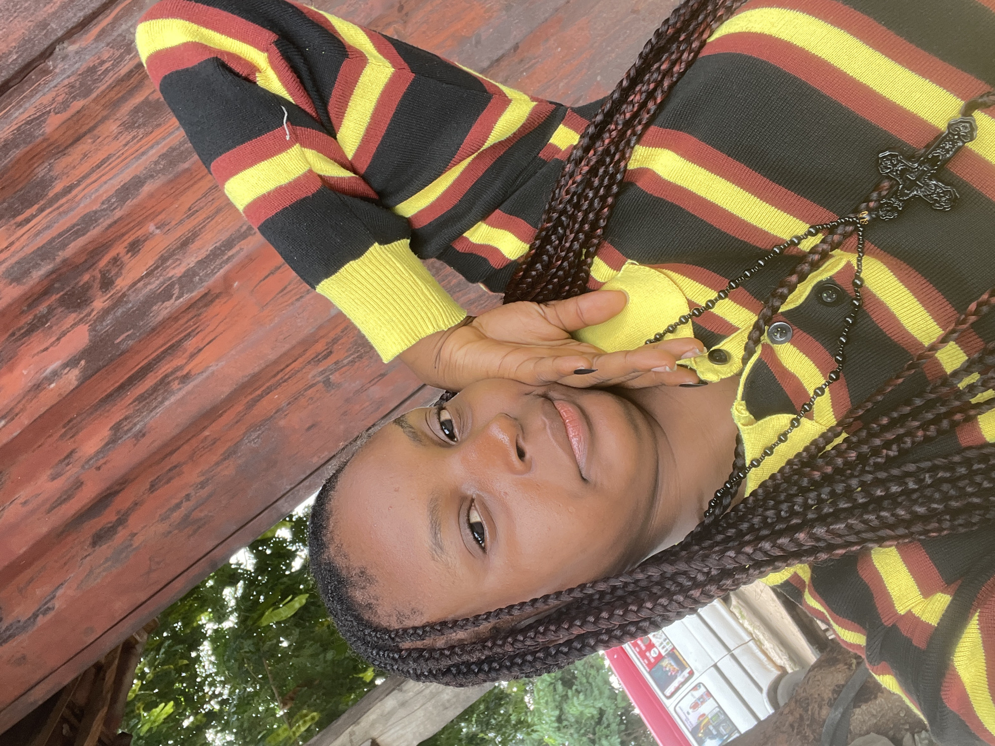 A young Black woman with braided hair is posing for a selfie in an outdoor setting. She is wearing a horizontally striped long-sleeved shirt in black, red, and yellow, and a dark necklace with a cross pendant. Her right arm is bent at the elbow, with her hand resting on her head. The background appears to be a weathered wooden structure and some greenery, with a white bus visible on the left. The lighting suggests it is daytime.