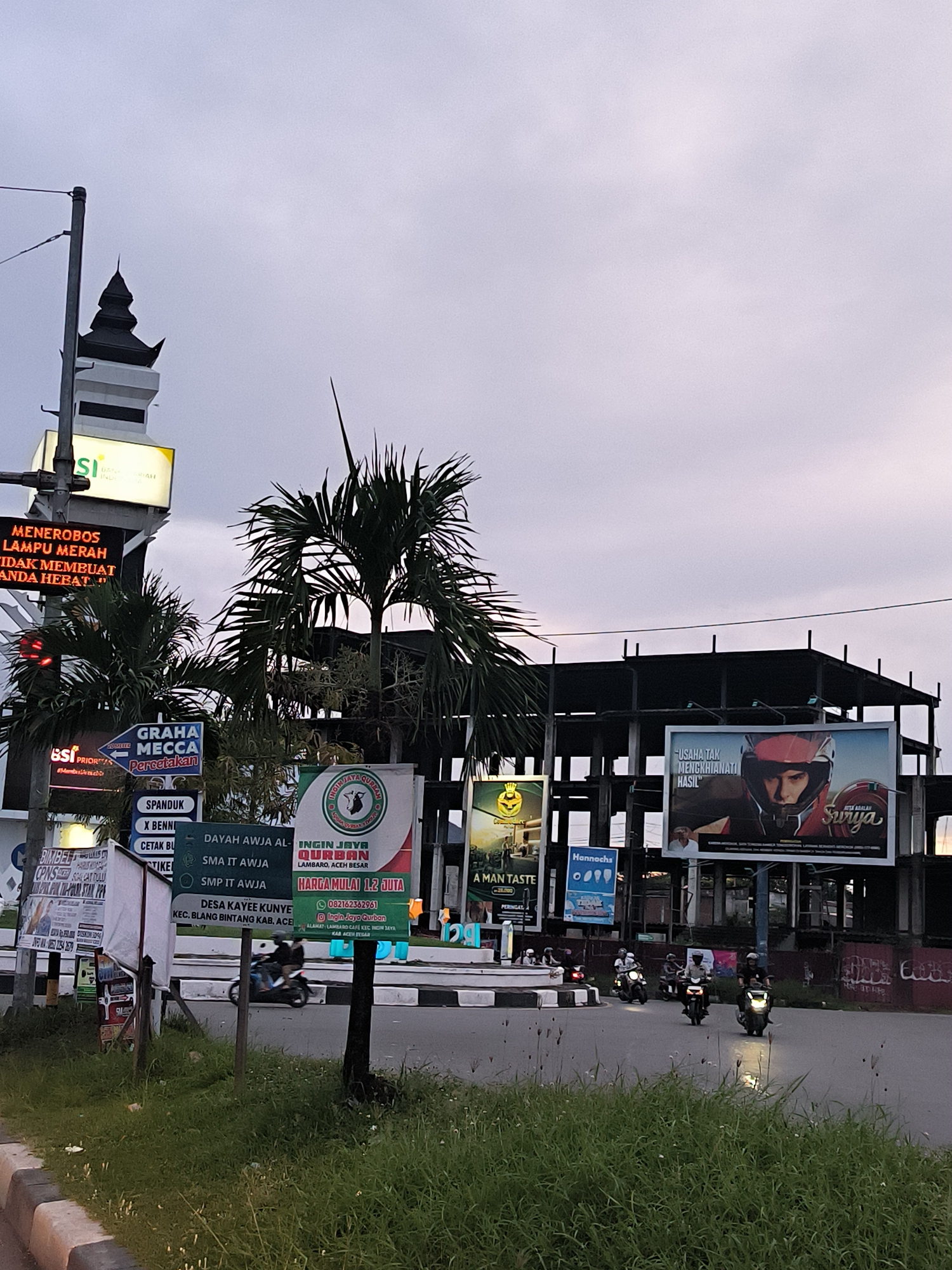 The image captures an evening street scene at an intersection in Lamprehlam Teungoh, Indonesia, under an overcast sky. In the foreground, a grassy roadside and median separate the viewer from the road. Several motorcyclists, wearing helmets, are visible on the street, some turning right around a small circular island.
To the left, a tall structure resembling a mosque minaret rises against the sky, alongside a prominent digital traffic light. The light is red, and a digital sign warns, "MENEROBOS LAMPU MERAH TIDAK MEMBUAT ANDA HEBAT!" (Running a red light does not make you great!). Below, various static signs advertise "GRAHA MECCA Percetakan" (Mecca Printing) and "BSI PRIOR."
In the mid-ground, numerous large billboards and smaller signs create a busy commercial environment. Lush palm trees are interspersed among the signs. One large green banner advertises "INGIN JAYA QURBAN" for "LAMBARO, ACEH BESAR," with prices starting from "HARGA MULAI 1.2 JUTA" and a contact number, also mentioning "DAYAH AWJA AL-," "SMA IT AWJA," "SMP IT AWJA," and "DESA KAYEE KUNYE KEC. BLANG BINTANG KAB. ACE." Another billboard features a person in a helmet with the text "USAHA TAK MENGKHIANATI HASIL" (Effort does not betray results) and the brand "Surya." Other visible brand names and services include "Hannochs" and "A MAN TASTE."
Behind the billboards, an unfinished or under-construction building with exposed steel or concrete frames is visible against the dim sky, adding to the urban/semi-urban setting. The overall impression is one of a busy commercial hub at dusk.