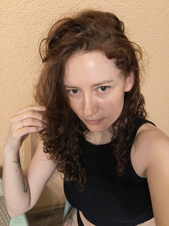 A female individual is depicted in a medium shot, looking directly at the camera. She has fair skin, dark brown eyes, and voluminous, auburn-colored curly hair, some strands of which are slightly disheveled. A small black hair clip is visible in her hair above her left temple. She wears a black ribbed sleeveless crop top, revealing a portion of her midsection and black undergarments below. Her left hand is raised, with fingers lightly touching the curls near her left ear; a thin, silver-colored ring is present on her left ring finger. A light-colored tattoo is visible on her left forearm, and a partial view of another tattoo, possibly a floral pattern, is seen on her upper right thigh. A segment of a light green or faded blue chair or stool is visible on the left side of the frame. The background is a plain, textured, light orange wall. The specific location within Saint Petersburg, Russia, cannot be determined from the visual evidence in the image.