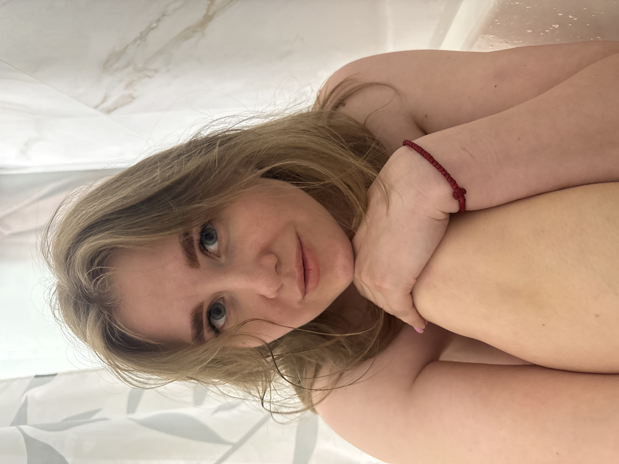 An image captures a young woman in a relaxed, reclined position, looking directly at the camera with a gentle, slightly smiling expression. She has blonde to light brown hair, which is slightly disheveled and frames her face. Her eyes are blue. She is wearing a red braided bracelet on her right wrist, and light pink nail polish is visible on one of her fingers.
The woman is shown from the chest up, with her arms bent and possibly supporting her head or resting on her body. Her skin appears fair. The background suggests an indoor setting, likely a bathroom or a tiled area. White surfaces dominate the background, including a prominent marble-patterned wall or surface with beige veining. On the far left, there are hints of a translucent shower curtain or partition, while the upper right corner shows what appear to be water droplets on a wet surface. The lighting is soft and even, highlighting her features without harsh shadows. No text is visible in the image.