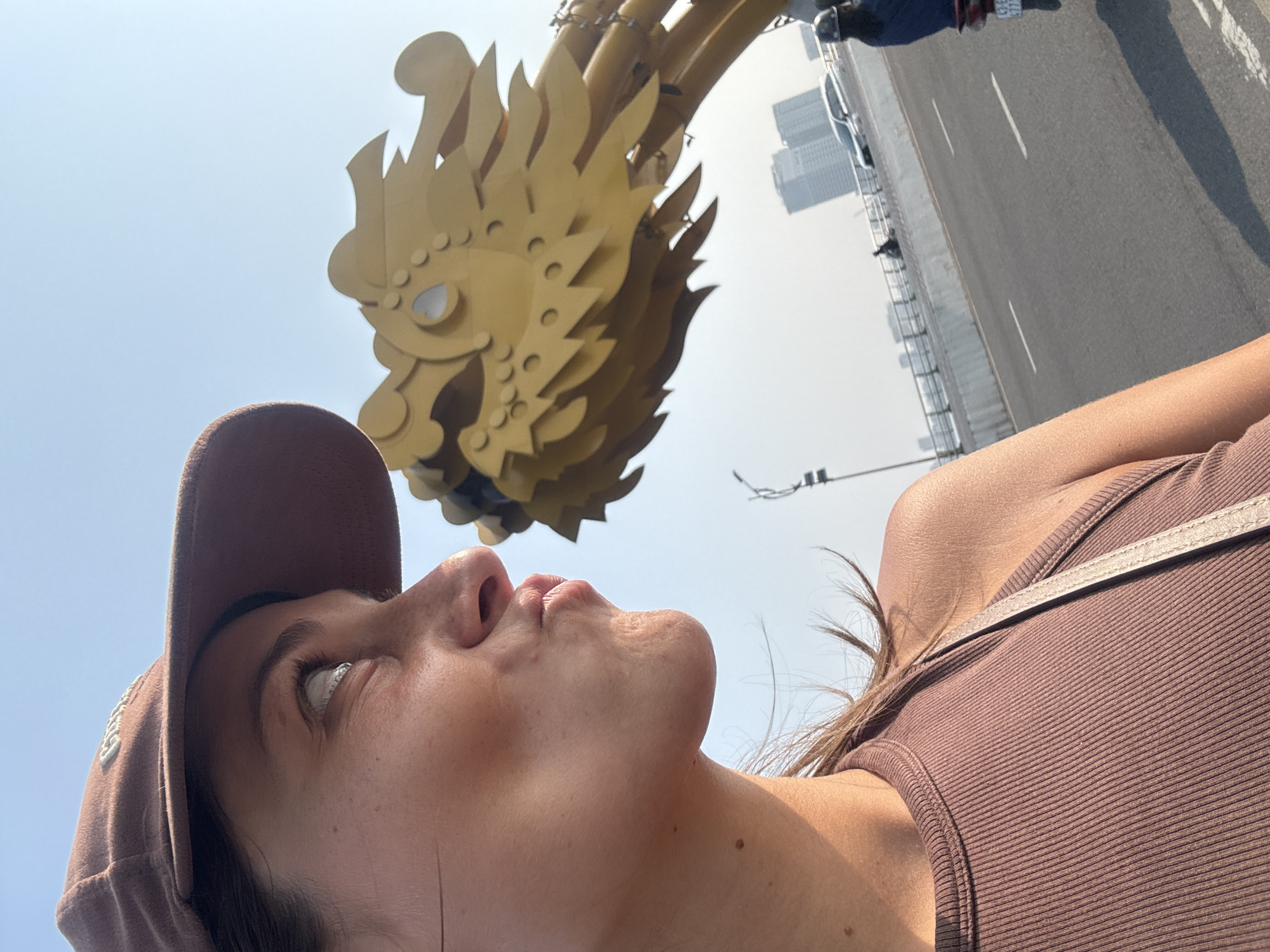 A woman, wearing a brown baseball cap and a brown ribbed tank top, looks up at the golden head of the dragon sculpture on the Dragon Bridge in Đà Nẵng, Vietnam. The bridge, including the road with cars and pedestrians, is visible in the background. The sky is light blue and hazy.