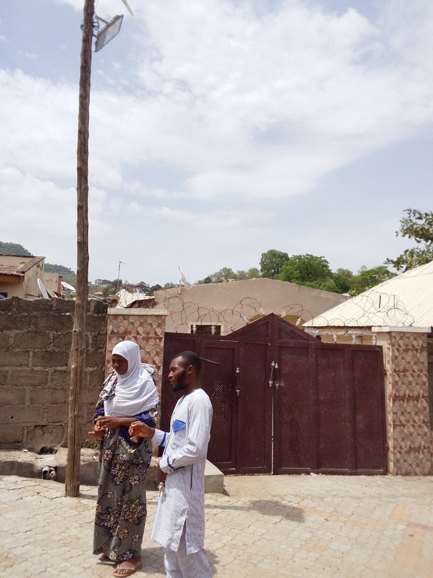 The image shows a man and a woman standing on a paved area next to a street in Lokoja, Nigeria. The woman is wearing a white headscarf and a long patterned dress. The man is wearing a striped long-sleeved shirt and pants. Behind them is a tall wooden pole with a modern light fixture attached. Behind the pole is a wall constructed of concrete blocks. To the right is a maroon metal gate, with a structure having a brown and white patterned design and razor wire on top. In the background, other buildings and trees are visible. The sky is cloudy.
