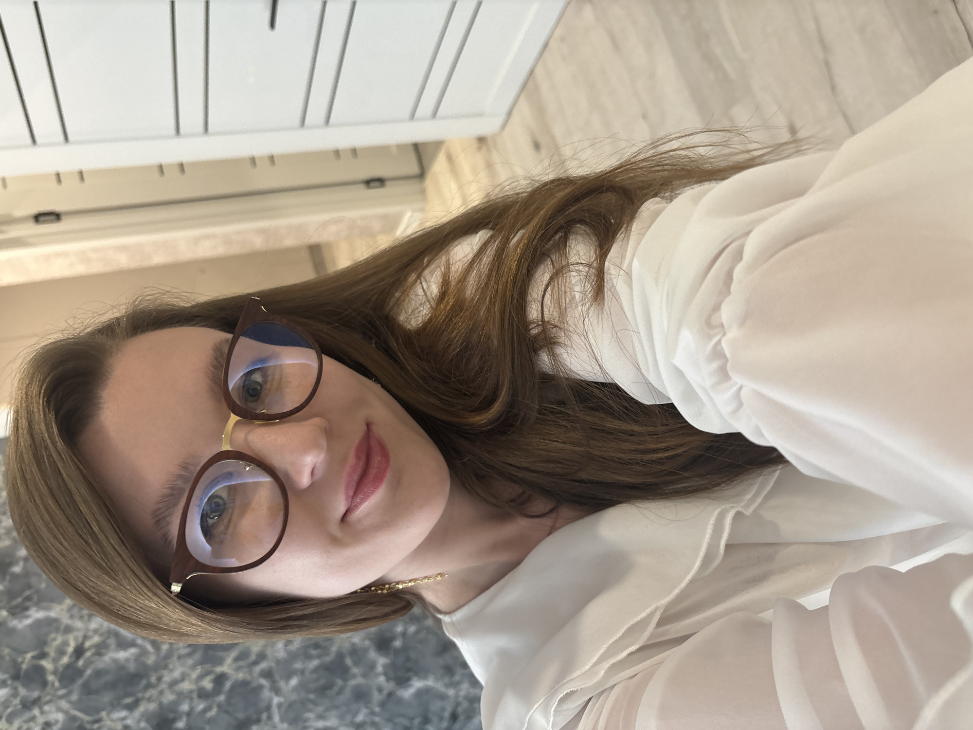 A fair-skinned woman with long brown hair is pictured from a slightly low angle, wearing brown-rimmed glasses. Her gaze is directed towards the camera, and she has a subtle smile. She is wearing a white, ruffled blouse and a gold necklace. The background is a blurred interior, showing a white cabinet with vertical lines on the top left and a marbled surface on the bottom left. There is no information available about the image location being in Izhevsk, Russia.