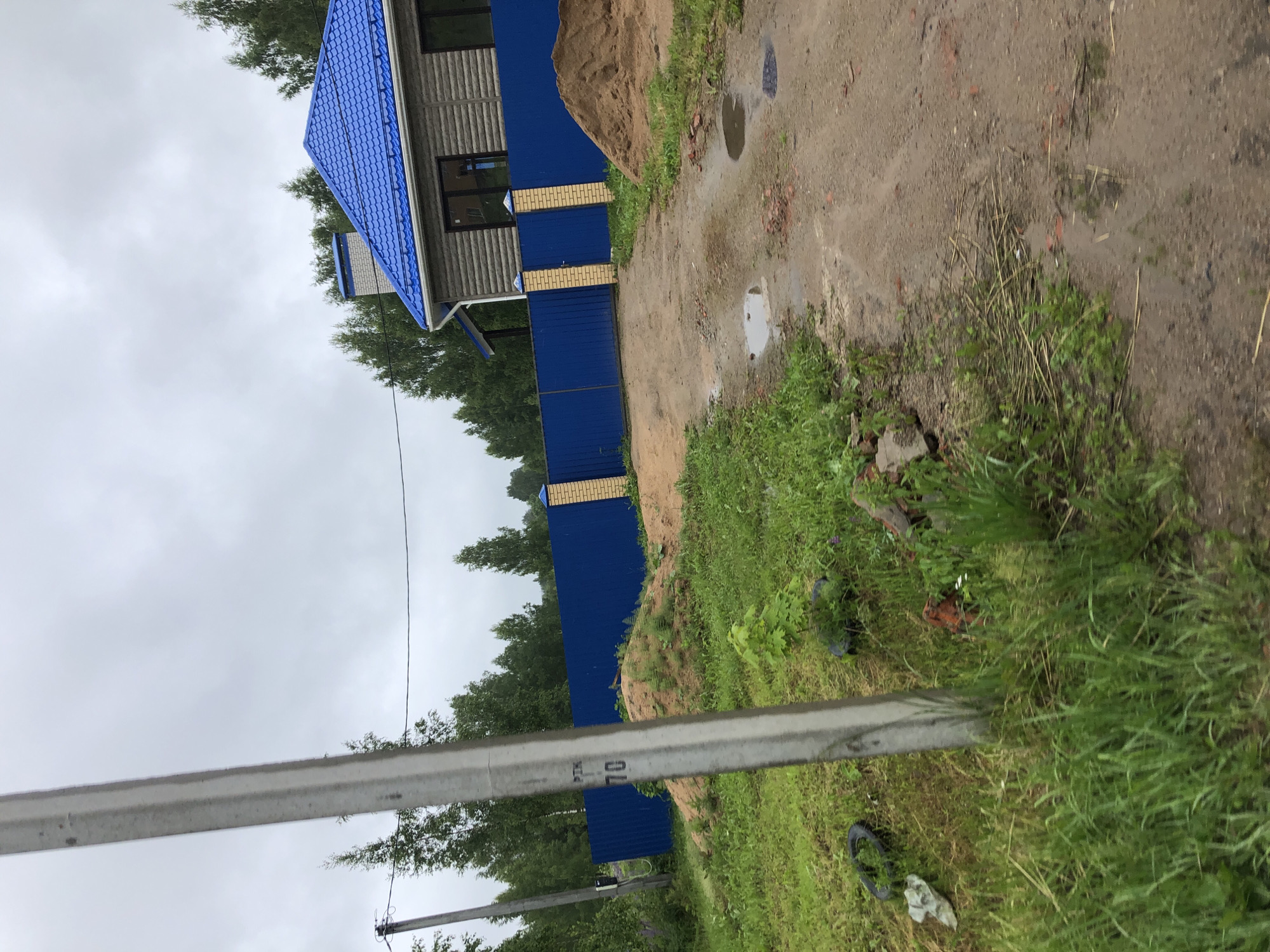 The image depicts an overcast day in an outdoor setting, likely a rural or semi-rural area near Safonovo, Russia, given the presence of a wooden-sided house with a blue metal roof and a matching blue metal fence. The property appears to be under construction or recently developed, with a large pile of sand on the right side of the dirt driveway. A concrete utility pole is prominent in the foreground on the left, with some faded markings visible on it. The ground is a mix of dirt and grass, with some puddles of water on the driveway suggesting recent rain. Green trees form a backdrop to the house and fence. There are no people or animals visible in the image. The overall impression is one of a work in progress in a somewhat undeveloped location.
