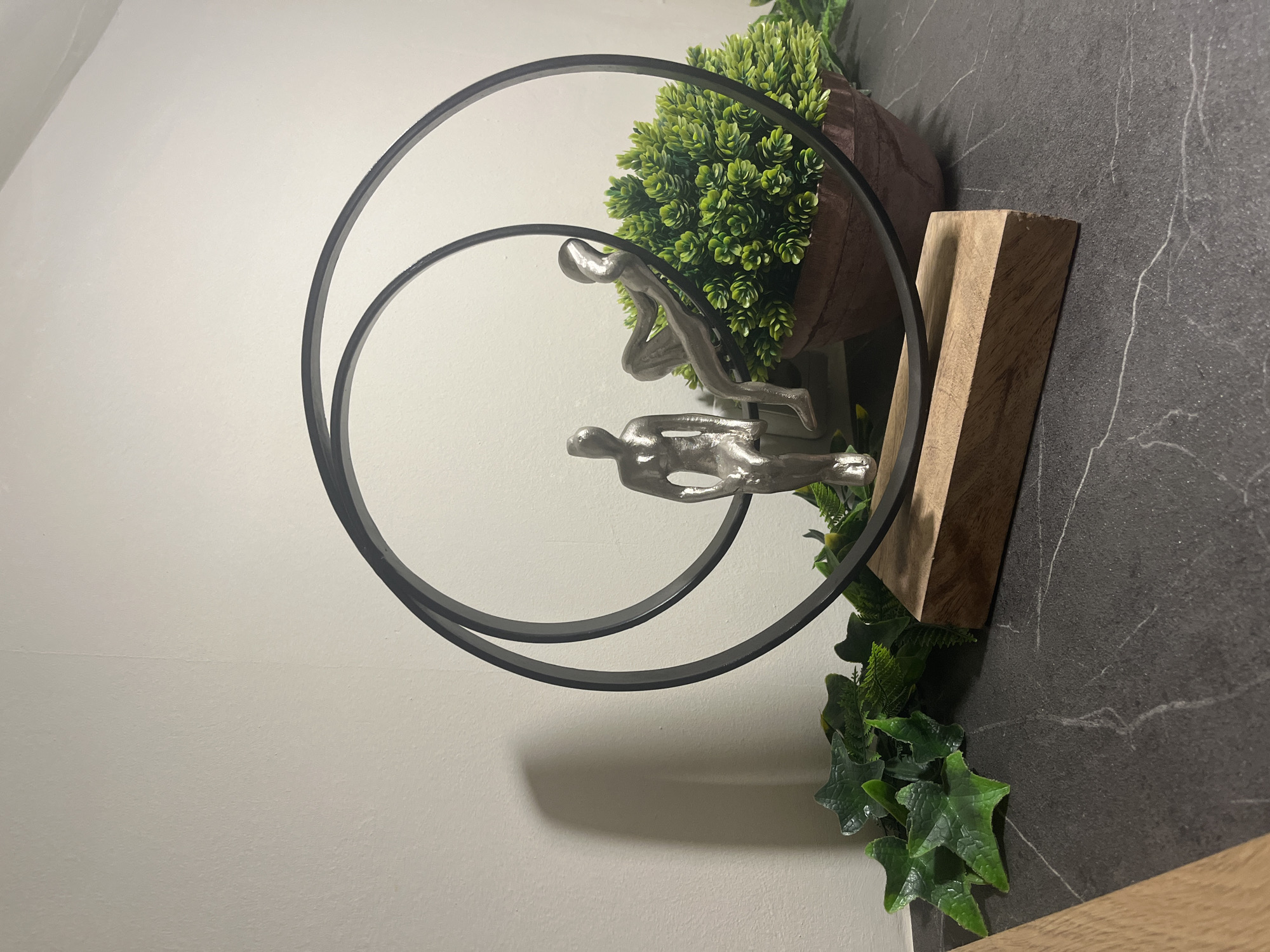 The image shows a decorative sculpture consisting of two silver figures in a black circular frame. The sculpture is partially surrounded by greenery, including a potted plant with small, bright green leaves, and ivy trailing down. A rectangular wooden block serves as part of the base. The sculpture is situated on a dark gray surface that appears to be a countertop with white veining, reminiscent of marble. The background consists of a plain white wall. The location is not discernible from the image, so it cannot be confirmed if it is located in Varna, Bulgaria.