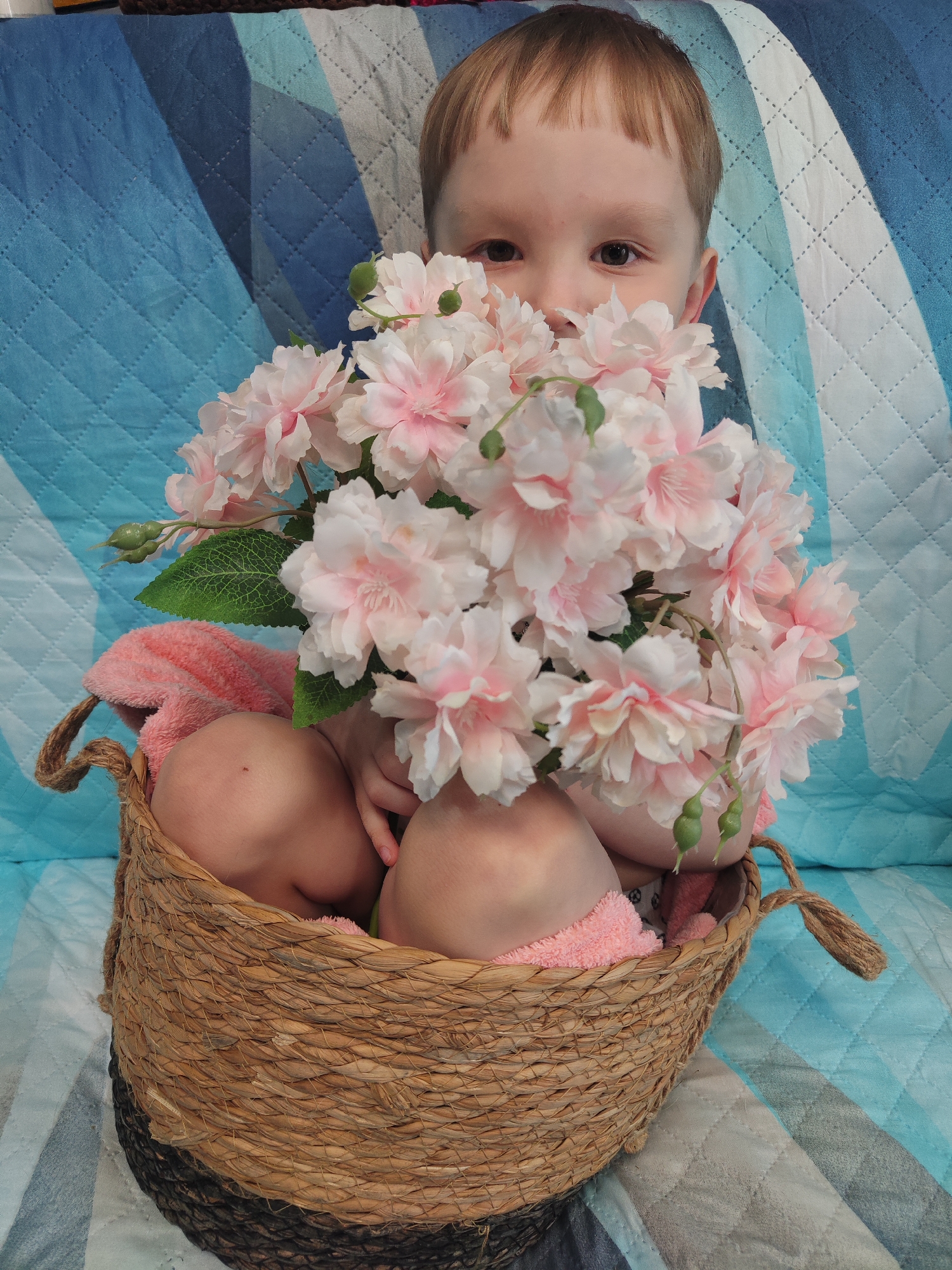 This close-up image, originating from Chaikovsky, Russia, features a young child playfully peeking from behind a large bouquet of artificial flowers. The child, with light brown hair and bangs, reveals only the upper half of their face, including wide, dark eyes and nose, looking directly at the camera with an expression of curiosity and innocence.
The dominant object in the foreground is a lush arrangement of pink and white faux flowers, possibly cherry blossoms or peonies, with visible green leaves and buds. These flowers are contained within a large, round basket woven from natural, light brown fibers, suggesting materials like seagrass or wicker. Inside the basket, partially visible beneath the child and flowers, is a soft, light pink towel or blanket. The child's body appears to be mostly nestled within this basket.
The background consists of a quilted fabric with alternating stripes of various shades of blue and white, indicating a domestic setting such as a bed or sofa. The lighting is even, suggesting an indoor environment. There is no visible text in the image. The photograph captures a sweet and tender moment, highlighting the child's interaction with the beautiful floral arrangement.