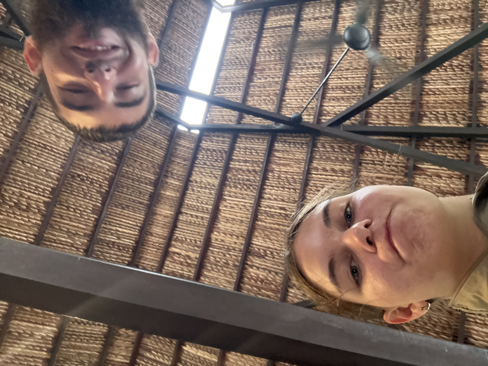 Two individuals are captured from a low-angle perspective, looking upwards towards a thatched roof structure. In the foreground, a young woman with blonde hair is looking directly at the camera with a slightly pursed expression. She is wearing a tan jacket and a small silver hoop earring in her left ear. Behind her and slightly to the right, a man with dark hair and a beard is also looking upwards, smiling with his eyes partially closed. The background consists of a rustic thatched ceiling supported by dark metal beams. The setting appears to be an open-air establishment, possibly a restaurant or resort common in coastal areas like Nha Trang, Vietnam, known for its tropical architecture and beachside resorts. The lighting suggests it might be daytime, with some natural light filtering through gaps in the roof.
