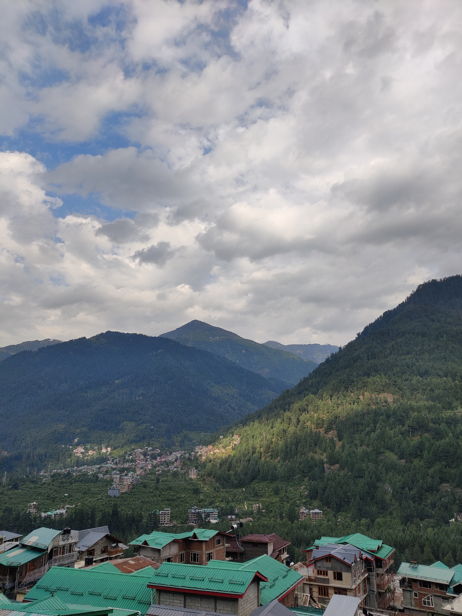 This is an aerial photograph of the town of Vashisht, India, taken on a partly cloudy day. The foreground shows a cluster of buildings with prominent green metal roofs, typical of the region. These buildings are multi-story structures, some appearing to be residential homes or guesthouses, with visible architectural details such as windows, balconies, and chimneys.
Beyond the foreground, the town extends up a verdant hillside, characterized by a dense forest of coniferous trees. Scattered amongst the trees are more buildings, forming a dense urban landscape on the slopes. The middle ground is dominated by these forested mountainsides, with varying shades of green indicating the depth and density of the foliage.
In the background, larger, more imposing mountains rise, their peaks shrouded in the diffused light of the overcast sky. One prominent mountain in the center appears to have a more rounded summit, while another to the right is steeper and more defined. Patches of blue sky are visible through gaps in the white and grey clouds that dominate the upper portion of the image, casting shadows and highlighting sections of the landscape. The overall impression is one of a mountain town nestled within a vast, forested landscape under a dramatic sky. There are no people visible in the frame.