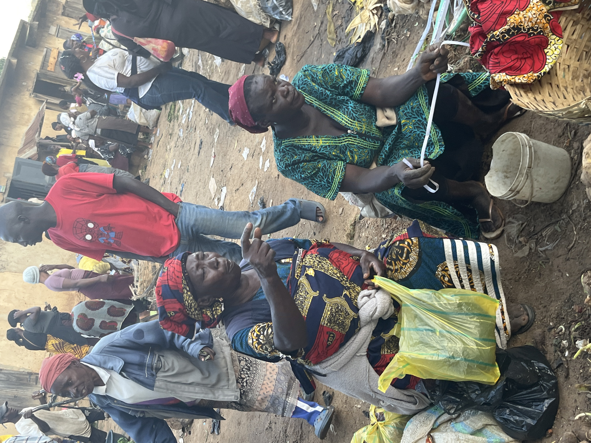 The image depicts a street market scene in Gura Top, Nigeria. In the foreground, two women are seated. The woman on the left, wearing a blue jacket and a red headscarf, is pointing to her right. She is dressed in traditional attire with a patterned skirt and a grey shawl. Next to her are yellow and black plastic bags. The woman on the right wears a green and yellow patterned dress and a red headscarf. Her hands are holding white plastic strips. In front of her are a white bucket and a woven basket containing fabric. In the background, other people are visible, some walking, and some standing. The ground is dirt with scattered debris. Buildings, appearing weathered, line the background. A man in a red t-shirt with a Spider-Man logo is also present in the mid-ground.