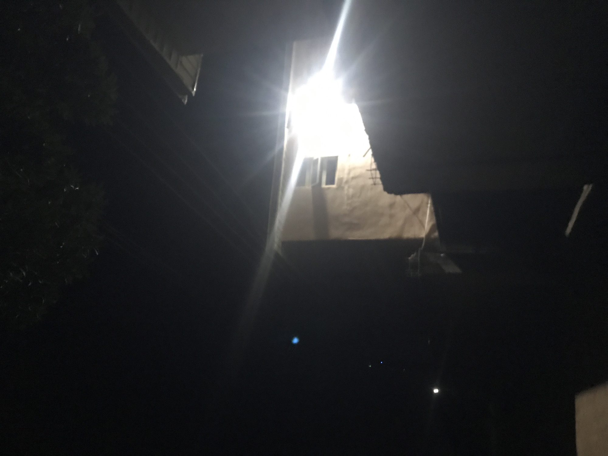 This image depicts a nighttime scene with a bright light emanating from a building. The building, likely a house, appears to be made of concrete and has at least one window. The light is so intense it causes lens flare, obscuring details of the interior. Some trees are visible in the upper portion of the frame, silhouetted against the dark sky. The ground is not clearly visible, but appears to be dark and possibly unpaved. The overall impression is of a dimly lit, residential area at night, potentially in a rural or semi-urban setting. There are no people visible in the image.