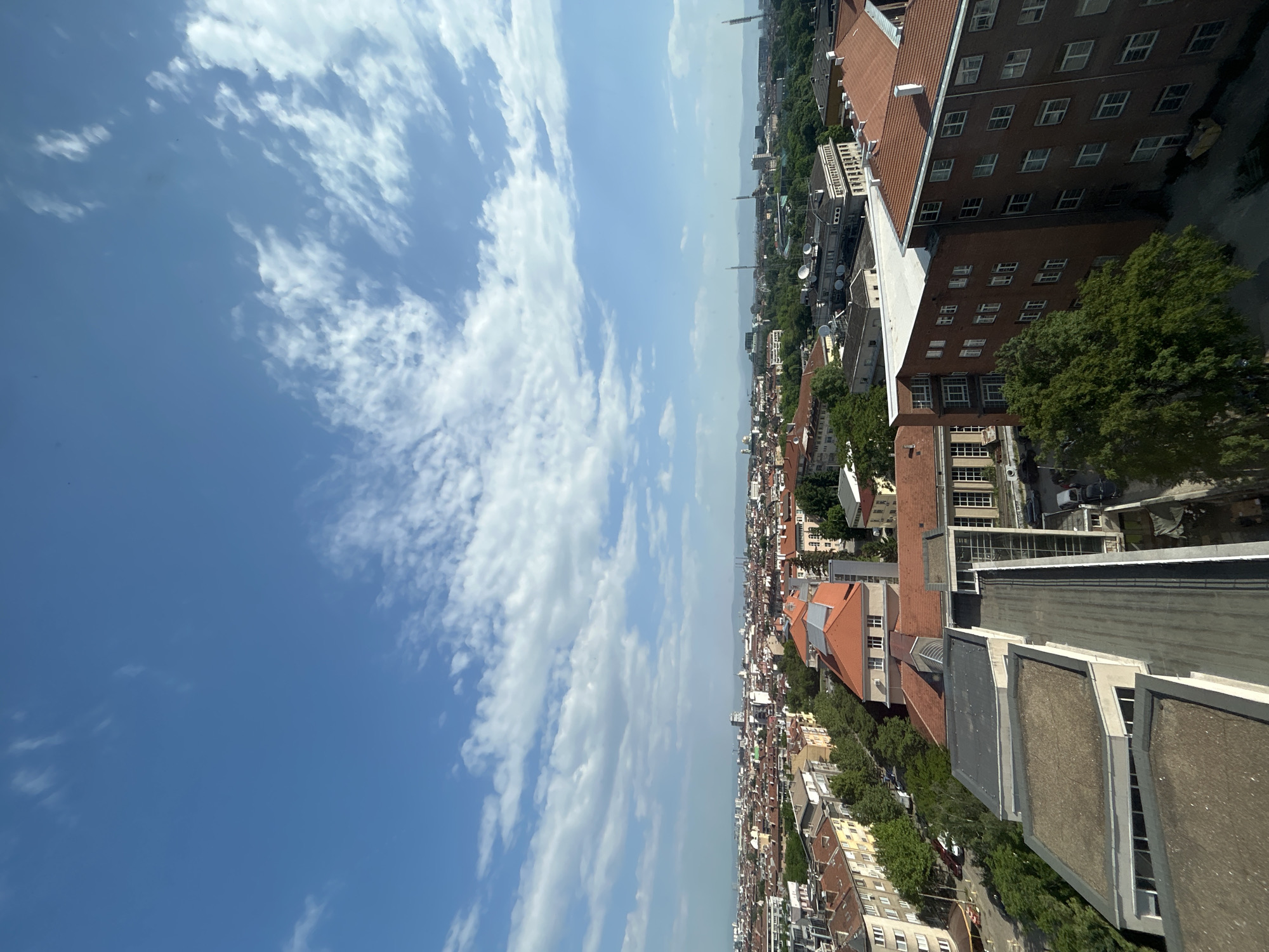 The image shows an aerial view of Sofia, Bulgaria, taken on a sunny day with scattered clouds. The cityscape consists primarily of red-roofed buildings of varying heights, interspersed with green trees. Tall buildings are visible towards the top of the image. In the distance, communication towers or antennas can be seen. The foreground shows parts of the building from where the photo was taken, with flat roofs and possible rooftop structures. No people are visible in the image.