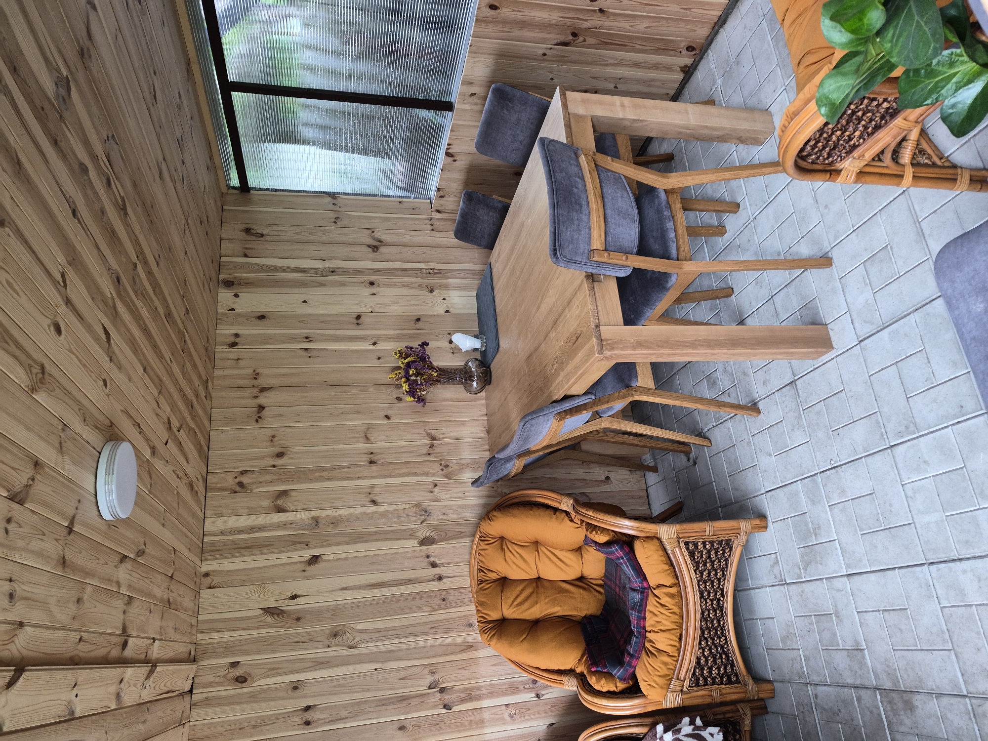 This image captures an interior view of a residential sunroom or enclosed porch in Hlevakha, Ukraine. The space features walls and a ceiling entirely clad in light-colored wooden panels, exhibiting visible grain and knots. The ceiling panels are oriented horizontally, while the wall panels are vertical. A single, circular white flush-mount light fixture is installed on the ceiling toward the upper left.
The floor is covered with light gray rectangular tiles laid in a herringbone pattern. A sturdy, rectangular wooden dining table occupies the central and right areas of the room. Four dining chairs with wooden frames and gray fabric upholstered seats and backs are arranged around the table. On the table's surface, a dark gray placemat holds a white tissue box, and a small glass vase contains a bouquet of dried purple and yellow flowers.
To the left of the dining table, a large wicker armchair is positioned, featuring a thick, tufted orange cushion. A dark plaid blanket is folded and rests on the armchair's seat. A segment of another wicker chair is visible in the lower left corner. A window with horizontal translucent slats or blinds is located in the upper right, revealing blurred green foliage beyond. A green leafy plant in a wicker planter is partially visible in the lower right corner. No individuals are present, and no activities are in progress.