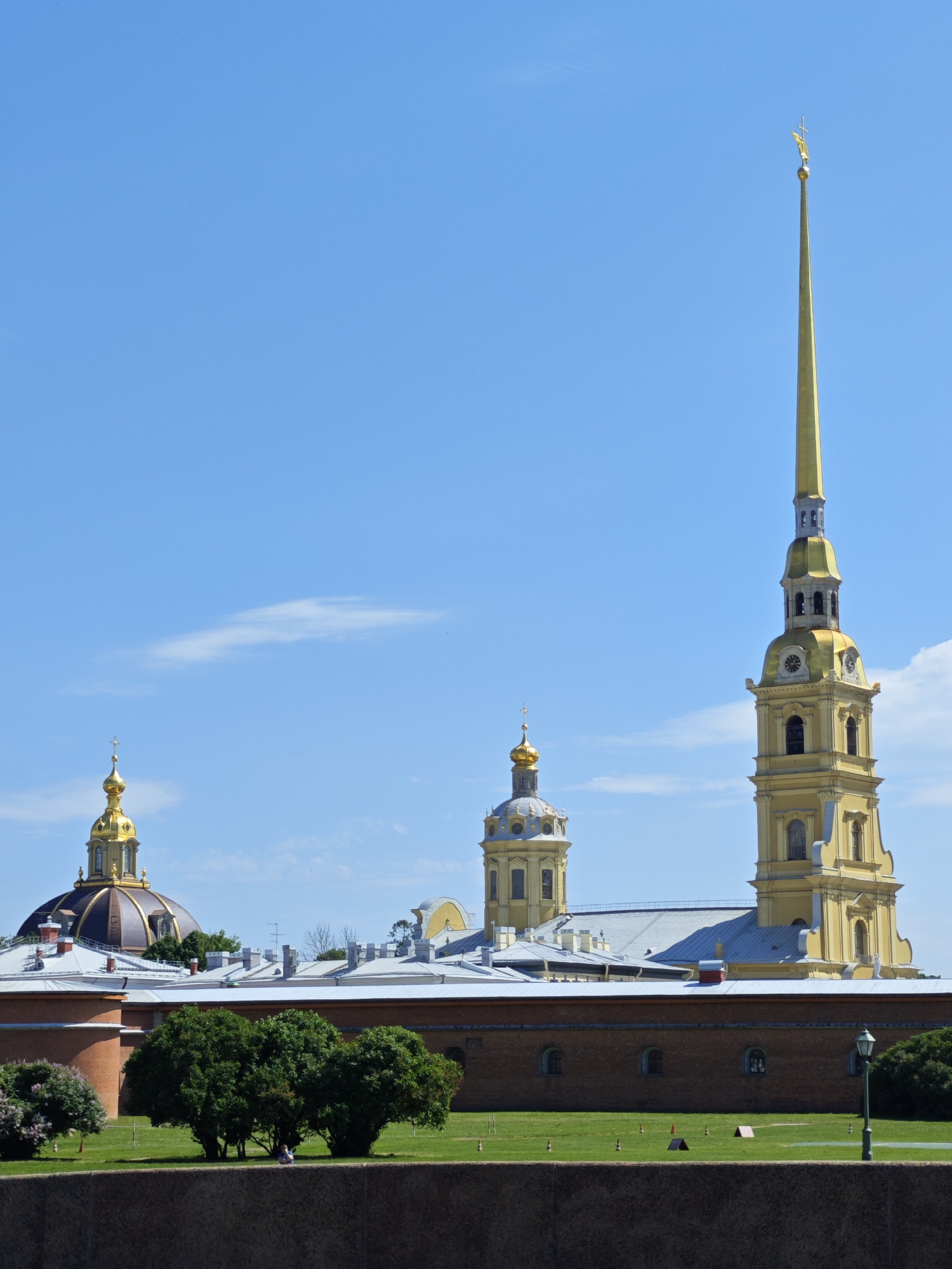 The image depicts a bright, sunny day with a clear blue sky and a few wispy clouds. In the background, the Peter and Paul Fortress in Saint Petersburg, Russia, stands prominently. The iconic Peter and Paul Cathedral, with its tall, golden spire topped by an angel, dominates the right side of the frame. To the left, a smaller golden-domed church with a cross also stands within the fortress complex. A long, low, brick wall forms the middle ground, with the roofs of various buildings visible behind it. In the foreground, a neatly mowed green lawn stretches across the scene, dotted with some small green bushes and a lilac bush on the far left. Two people are seated on the grass, appearing to be enjoying the day. A green lamppost is visible on the right side of the lawn. The overall impression is one of historical grandeur and a peaceful, pleasant atmosphere.